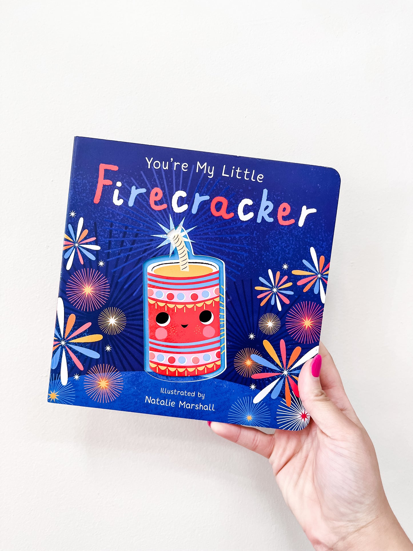 A hand with pink nail polish holds the My Little Firecracker Book by Tiny Tot Bundles, featuring a smiling firecracker and colorful fireworks on a blue cover.