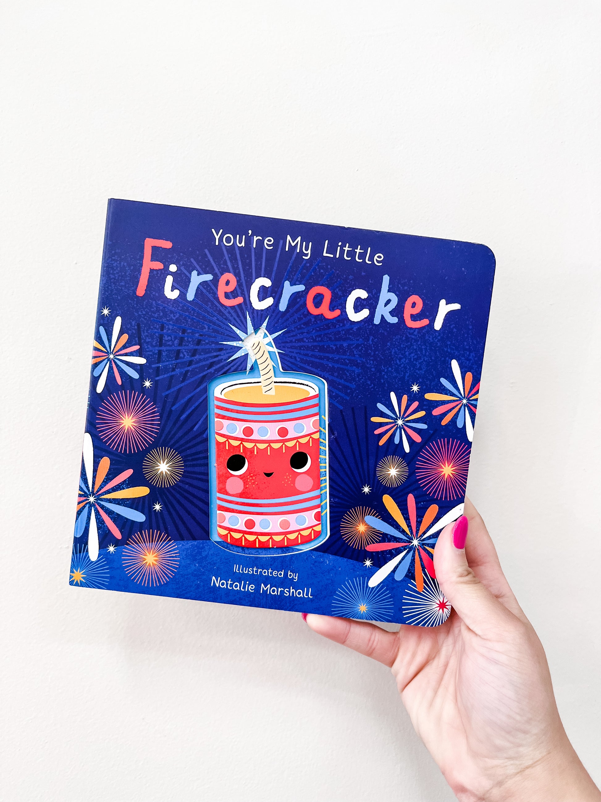 A hand with pink nail polish holds the My Little Firecracker Book by Tiny Tot Bundles, featuring a smiling firecracker and colorful fireworks on a blue cover.