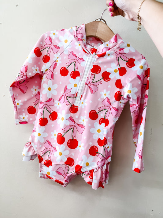 A hand displays the Tiny Tot Bundles Strawberry Daisy Swimsuit—a pink long-sleeve baby swimsuit with a zipper, decorated with red strawberries, white daisies, and pink bows—hanging on a wooden hanger against a light background.