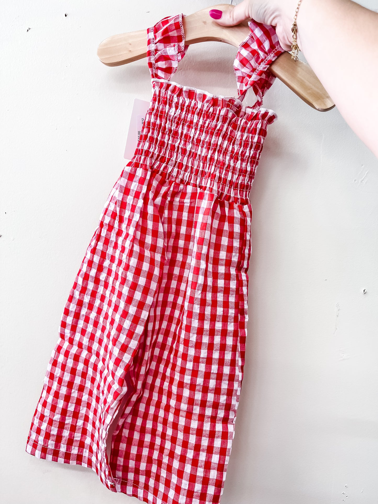 A hand holds the Tiny Tot Bundles Red Gingham Romper, a smocked sleeveless children’s outfit with a red and white check pattern, displayed on a wooden hanger against a plain white wall.