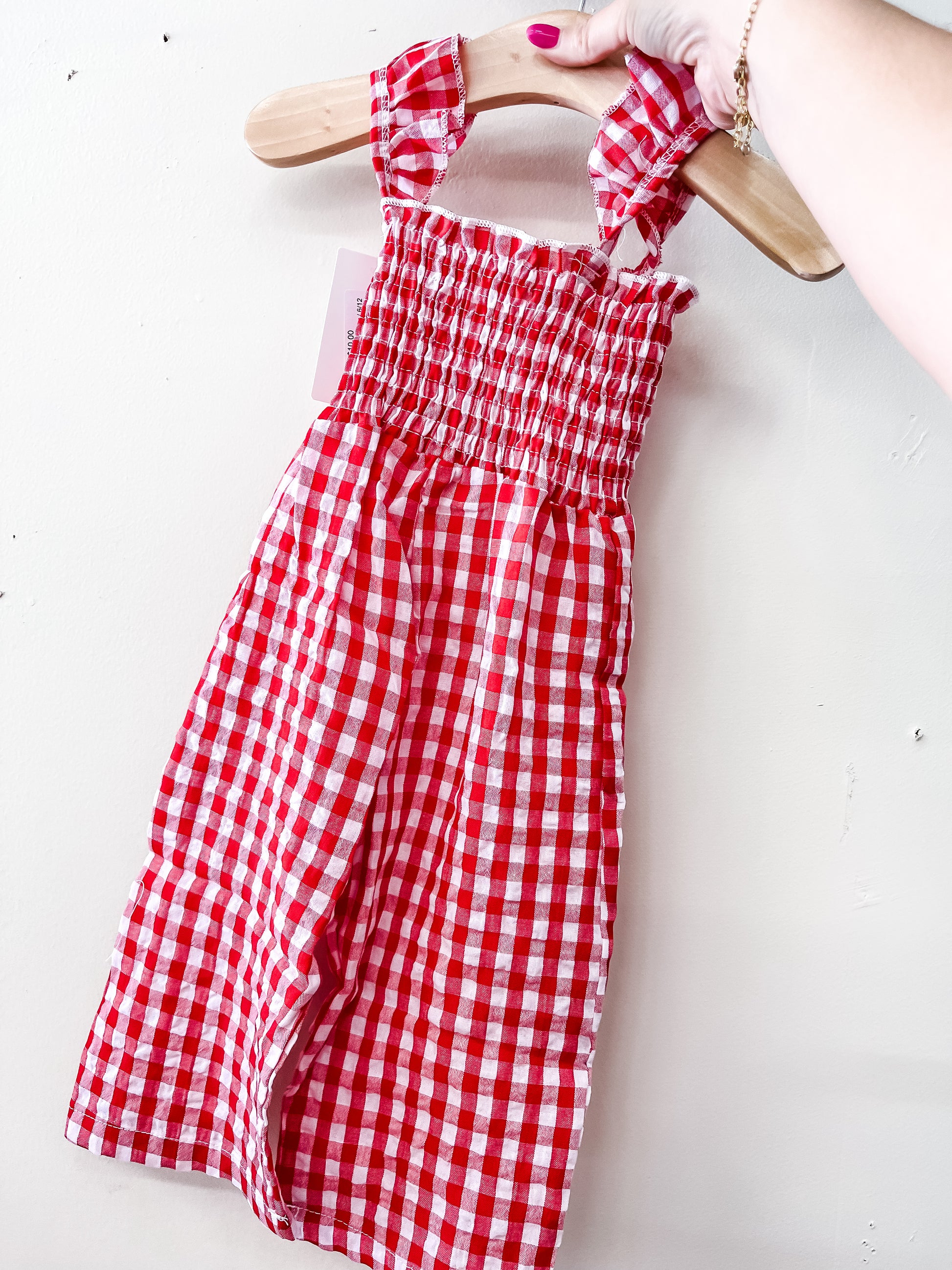 A hand holds the Tiny Tot Bundles Red Gingham Romper, a smocked sleeveless children’s outfit with a red and white check pattern, displayed on a wooden hanger against a plain white wall.