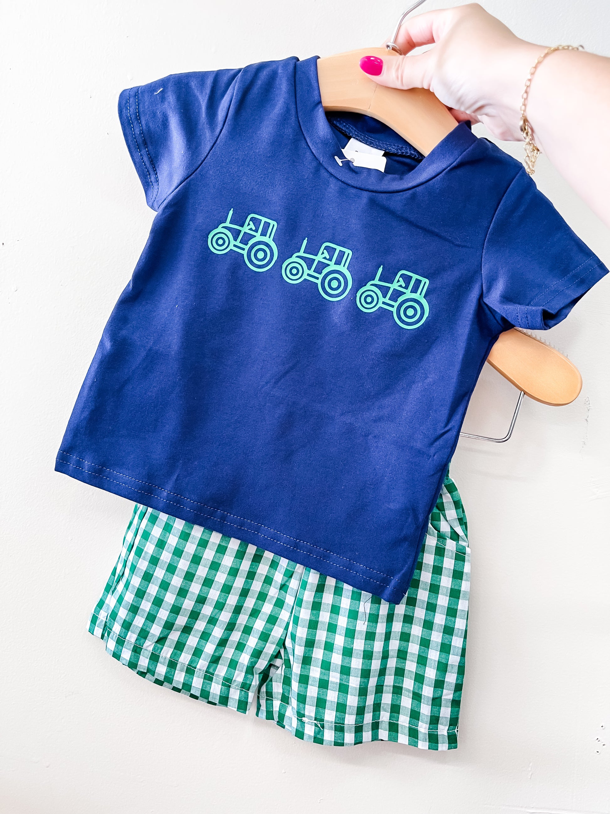 A hand holds the Tiny Tot Bundles Green & Blue Tractor Gingham Set: a navy t-shirt with three green tractors and matching green-and-white checkered shorts.
