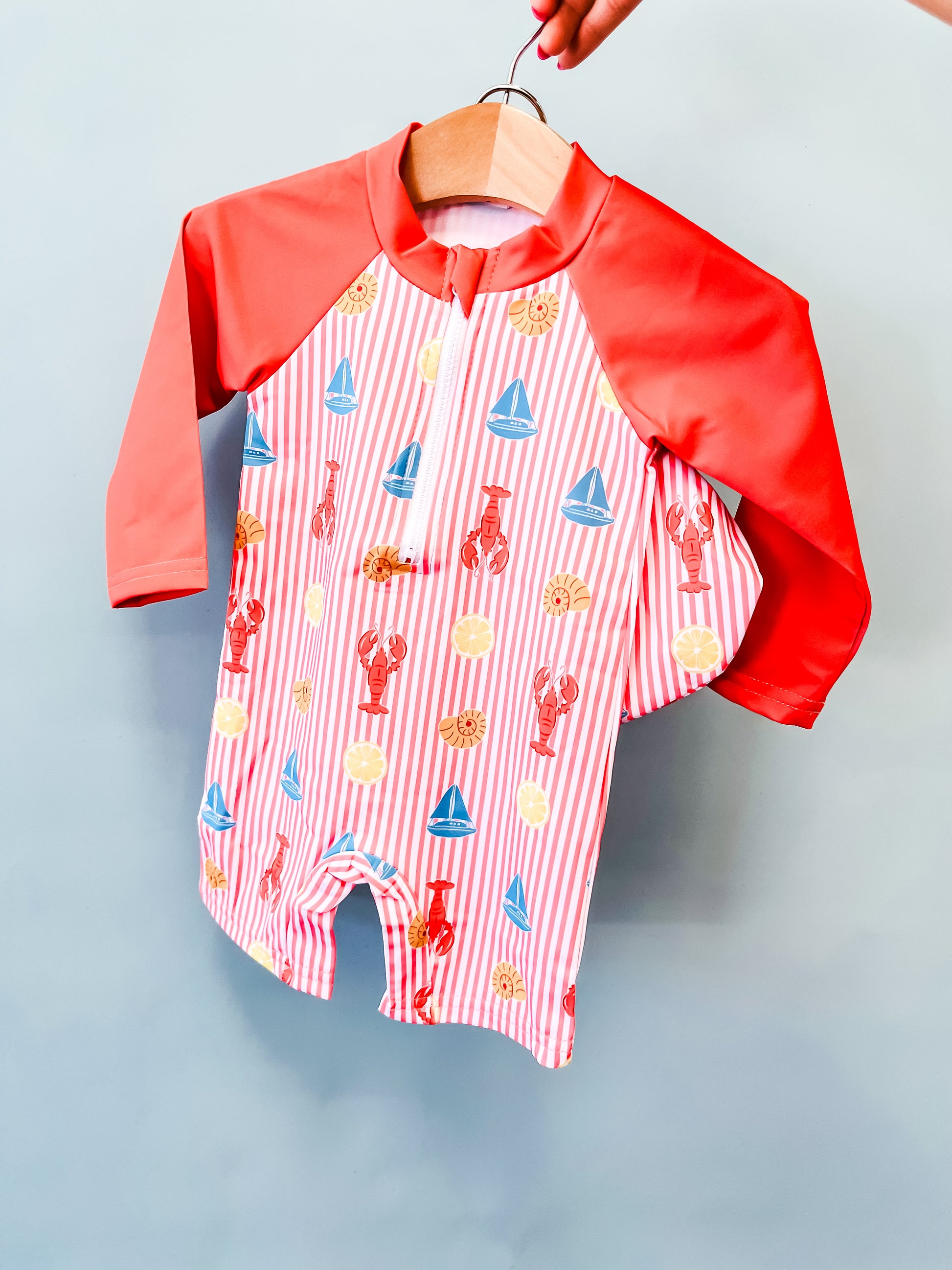The Tiny Tot Bundles Lobster Lagoon Swimsuit features red sleeves and pink-and-white stripes, adorned with playful lobster, sun, and blue sailboat prints. Displayed on a wooden hanger against a light blue background.