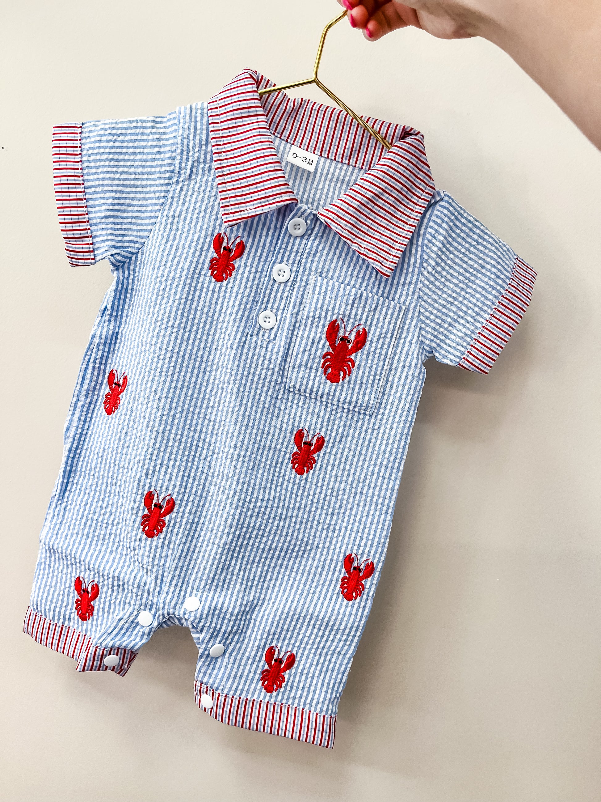 The Lobster Dock Romper by Tiny Tot Bundles features blue and white stripes, red lobster prints, red and white striped collar and cuffs, and white buttons, displayed on a small gold hanger.