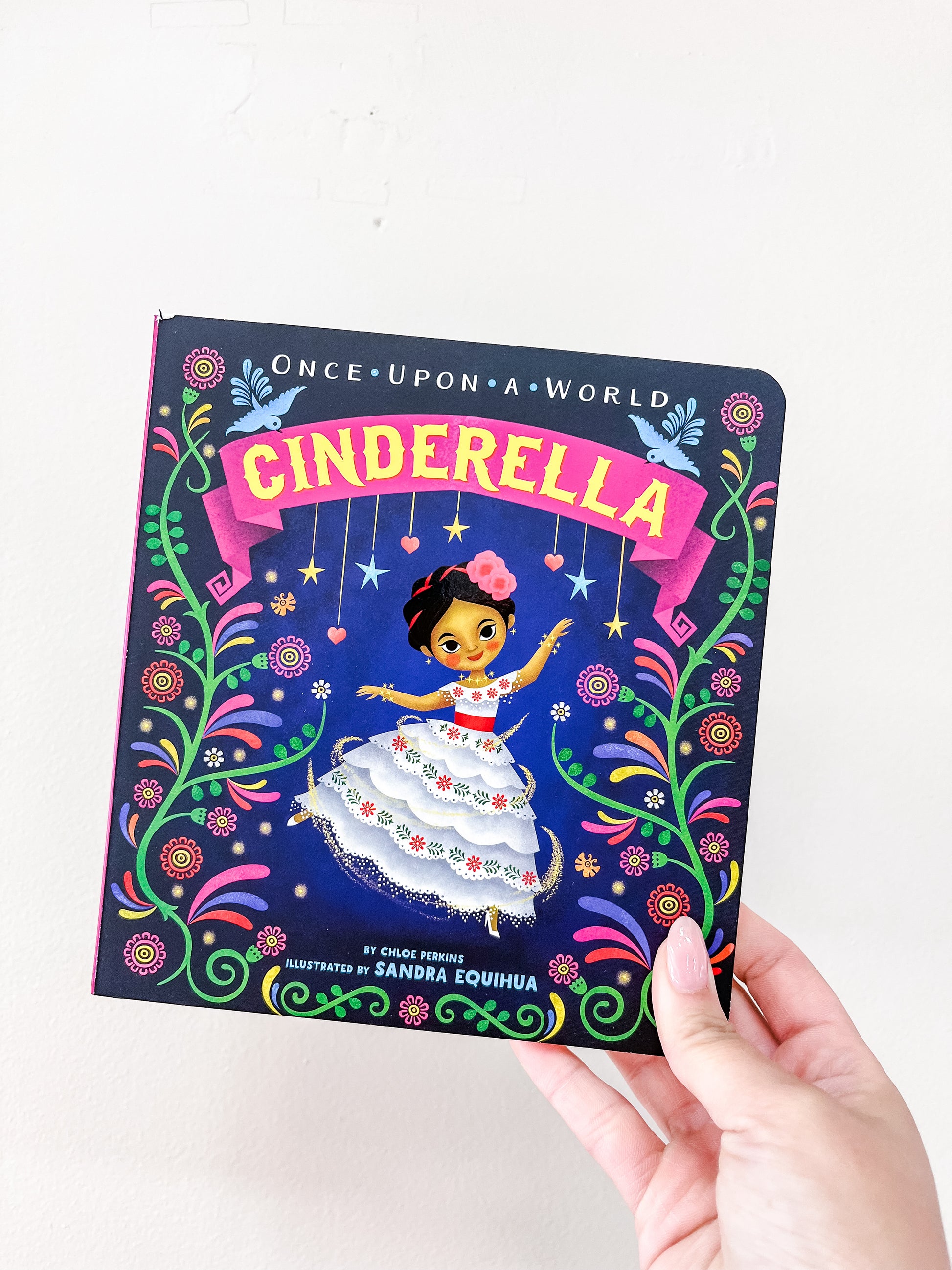 A hand holds the Cinderella Book by Simon & Schuster, featuring an illustrated girl in a white dress with flowers on a blue background with vibrant floral designs and the text "Once Upon a World" above.