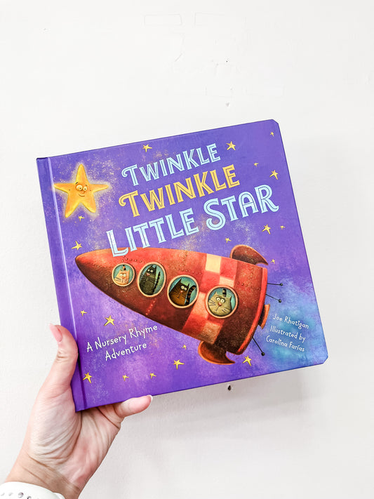 A hand holds the Twinkle, Twinkle Little Star Book by Simon & Schuster, featuring a purple cover with a rocket ship, animal characters, and a smiling yellow star.