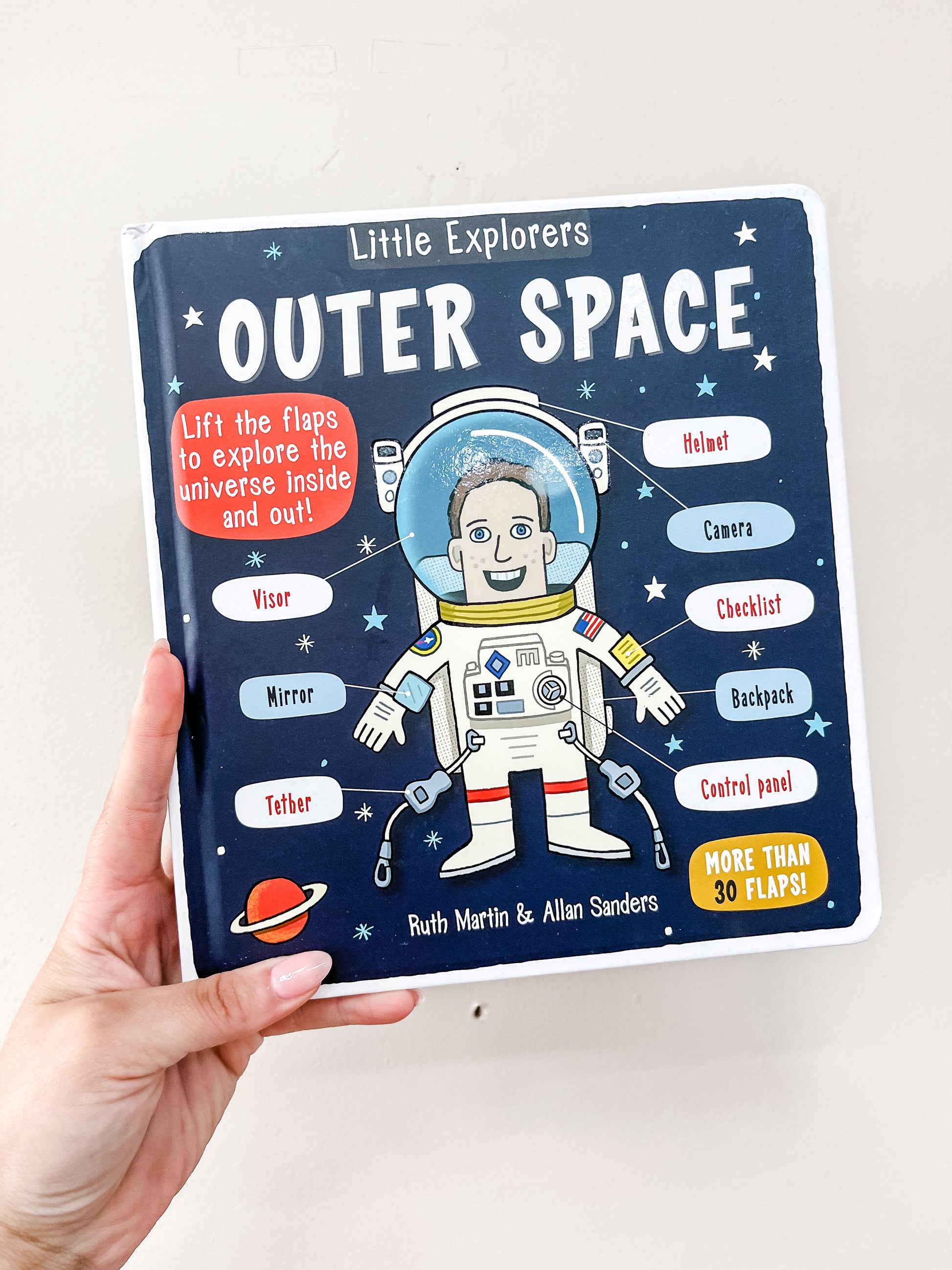 A hand holds the Simon & Schuster Little Explorers: Outer Space Book, featuring a cartoon astronaut and labeled space items on the cover, with text showcasing its interactive flaps.