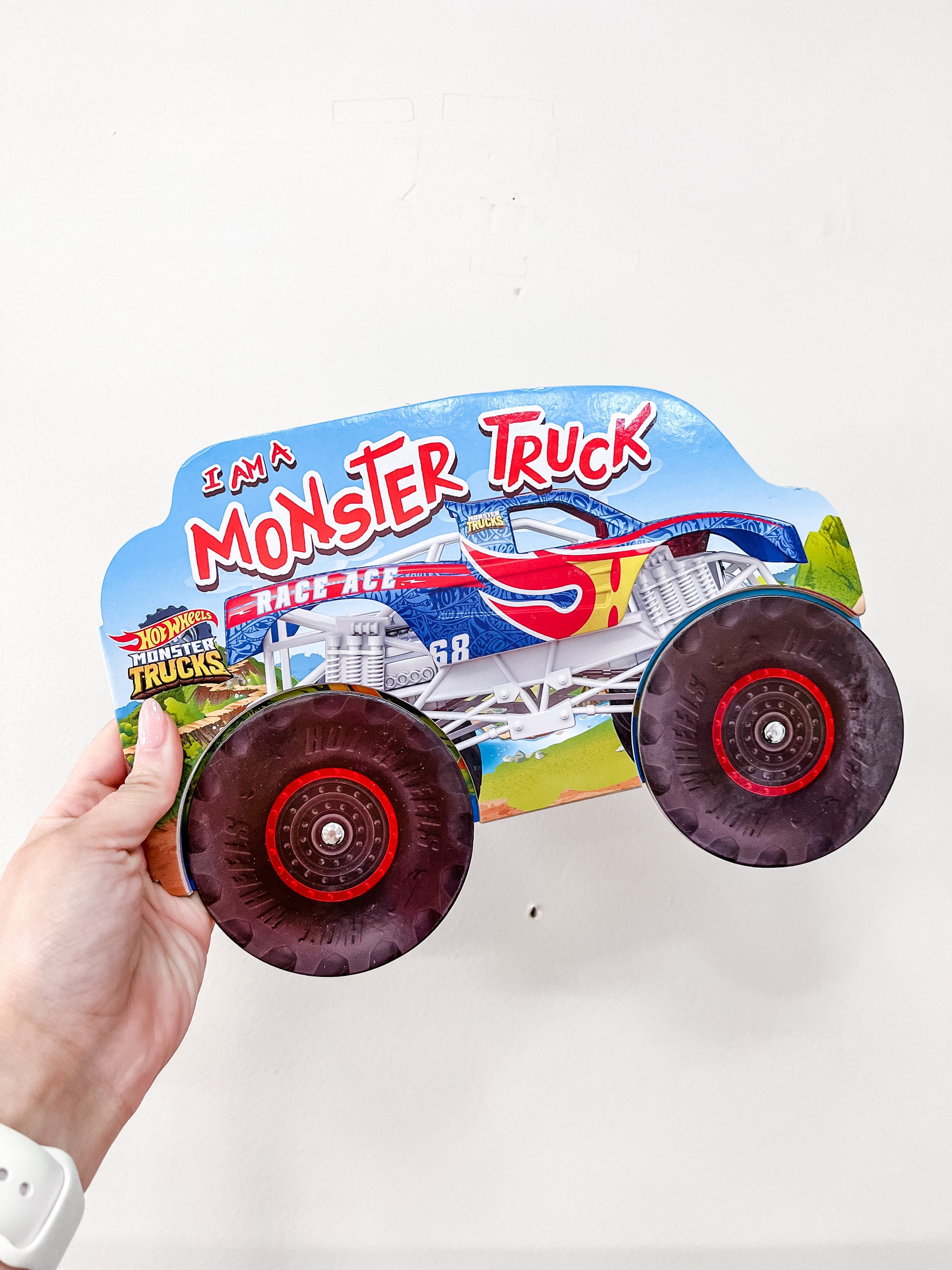 A hand holds the Simon & Schuster “I Am A Monster Truck Book,” a children’s book shaped like a monster truck with large wheels, against a plain white background.