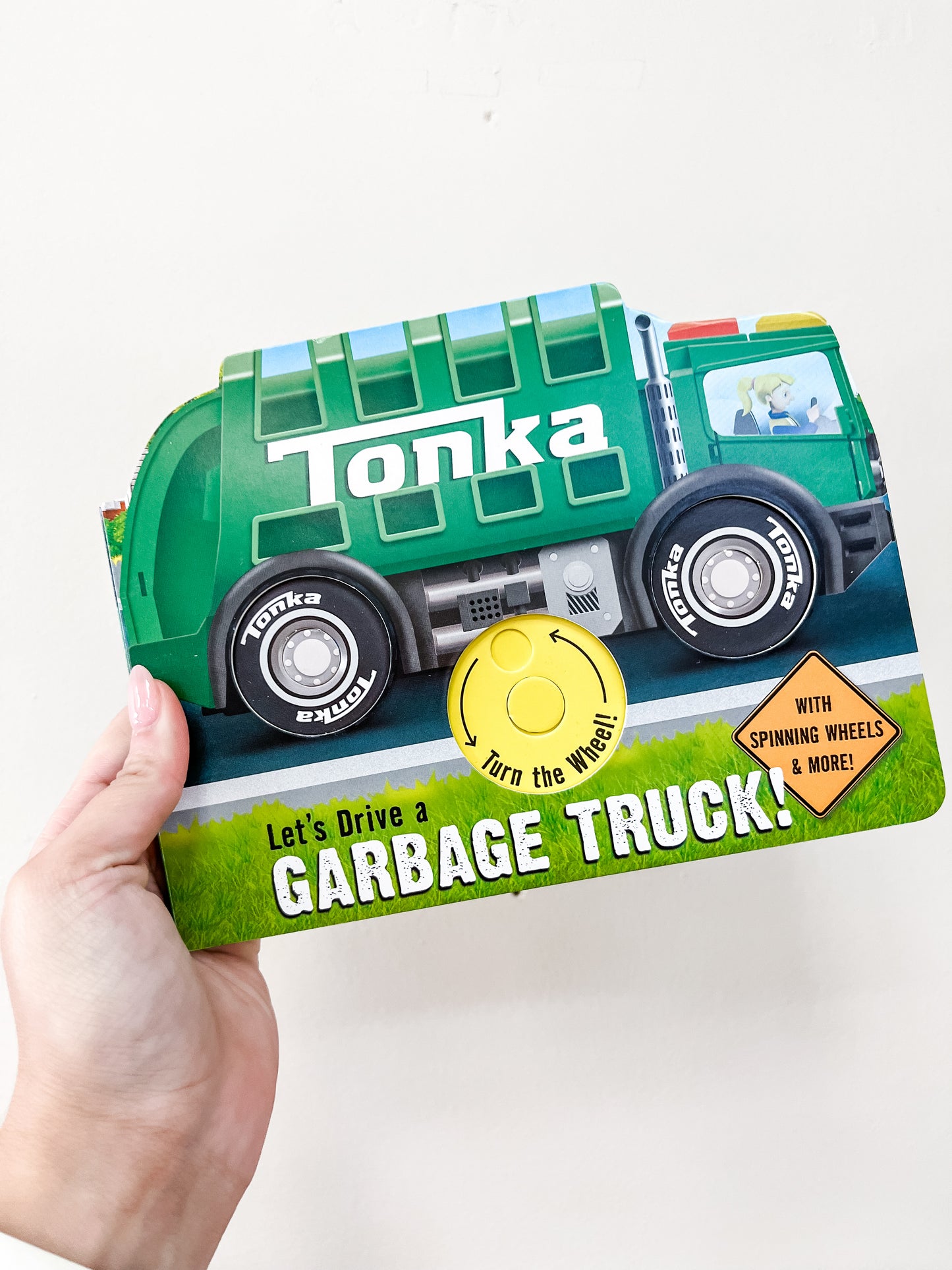 A hand holds the Simon & Schuster Tonka: Let's Drive A Garbage Truck Book, a children's book shaped like a green Tonka garbage truck with spinning wheels and a yellow "turn the wheel!" circle on its cover.