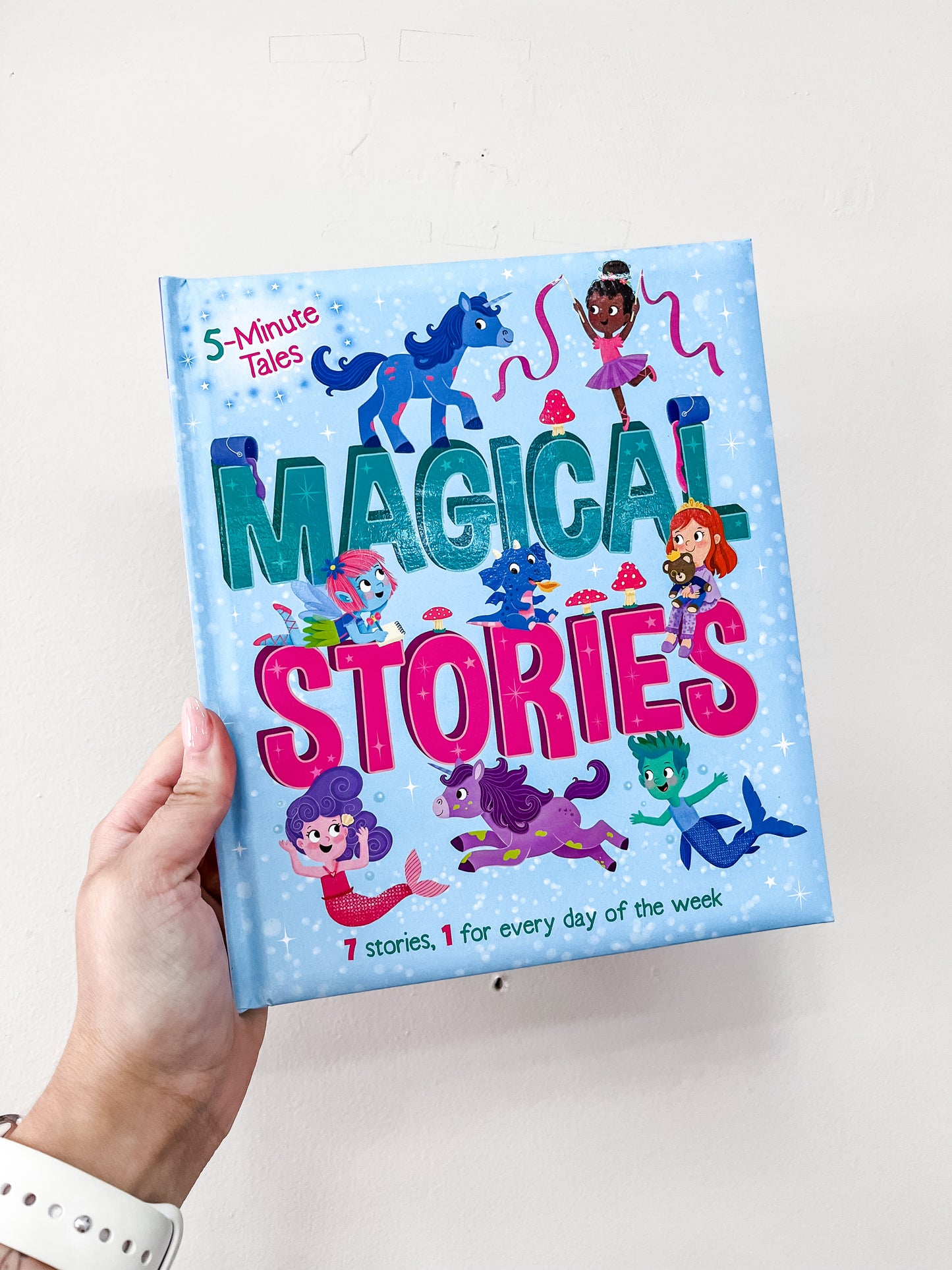 A hand holds Simon & Schuster’s 5 Minute Tales: Magical Stories Book, featuring a blue, starry cover with colorful illustrations of fairies, unicorns, and magical creatures.