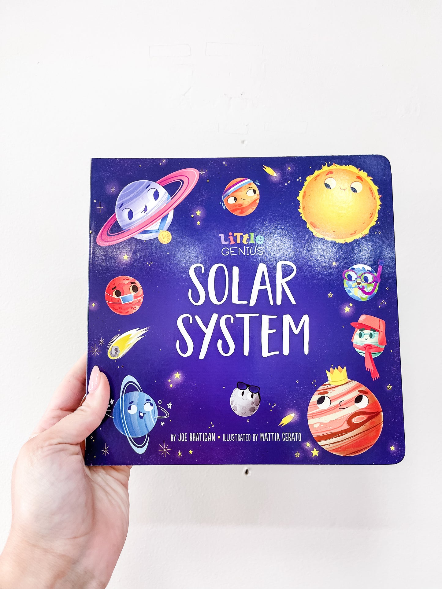 A hand holds the Little Genius Solar System Book by Simon & Schuster, a children’s board book featuring cartoon planets and the sun with faces on a dark blue, starry cover.