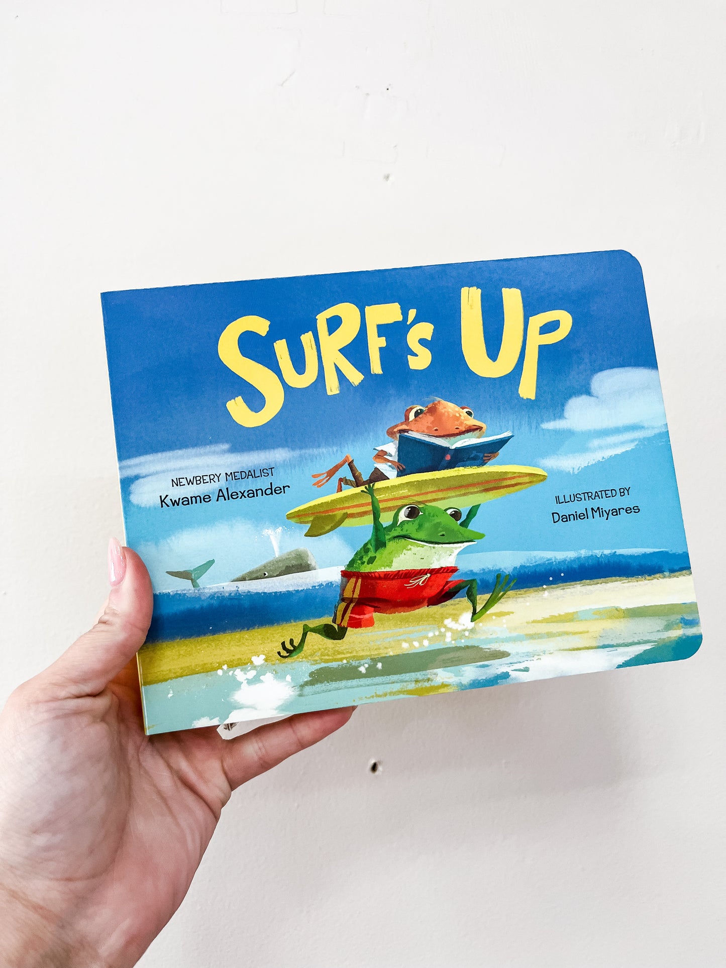 A hand holds the Surf's Up Book by Simon & Schuster. The cover features two frogs at the beach—one carrying a surfboard, the other riding on top—with water and clouds in the background.