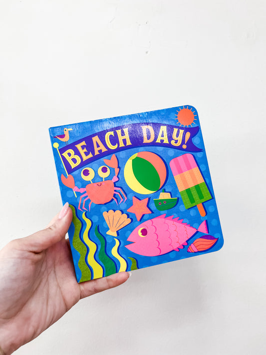A hand holds the Beach Day Book by Simon & Schuster, a colorful children's book featuring playful illustrations of a crab, fish, seashell, beach ball, popsicle, and sun on a vibrant blue cover.