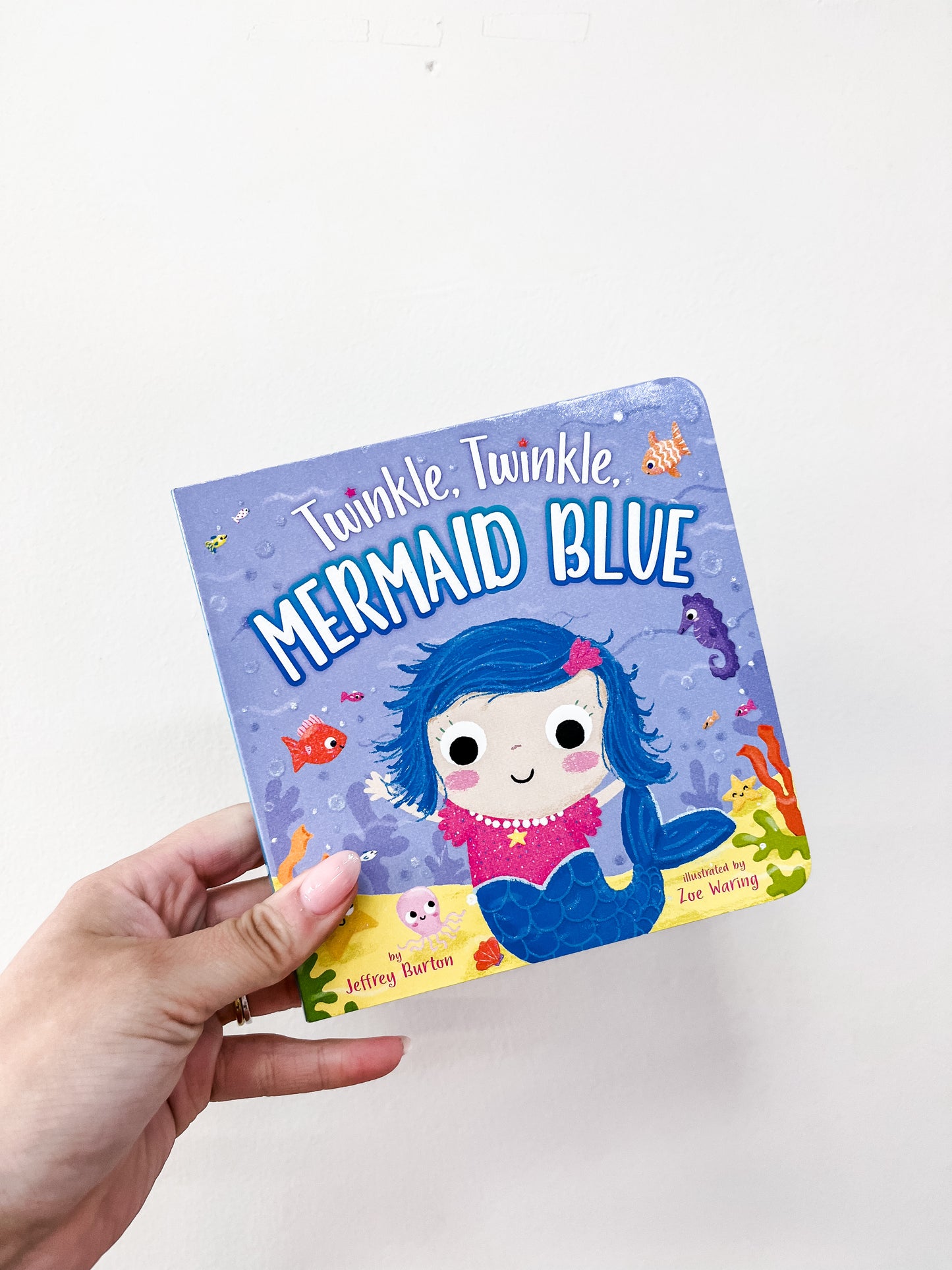 A hand holds the Simon & Schuster Twinkle, Twinkle, Mermaid Blue Book, featuring an illustrated mermaid with colorful sea creatures and coral on a light purple cover.