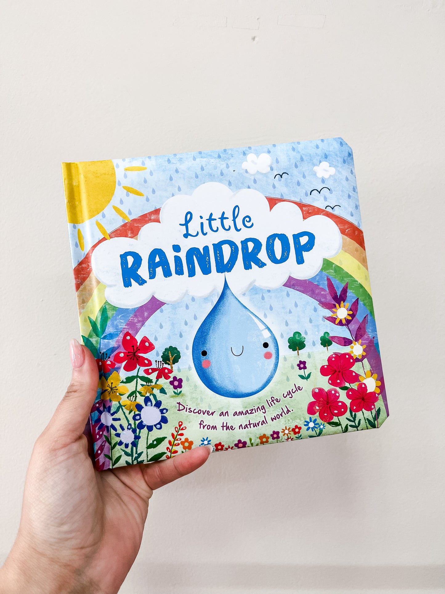 A hand holds the Little Raindrop Book by Simon & Schuster, with an illustrated cover showing a smiling raindrop, rainbow, sun, clouds, and colorful flowers.