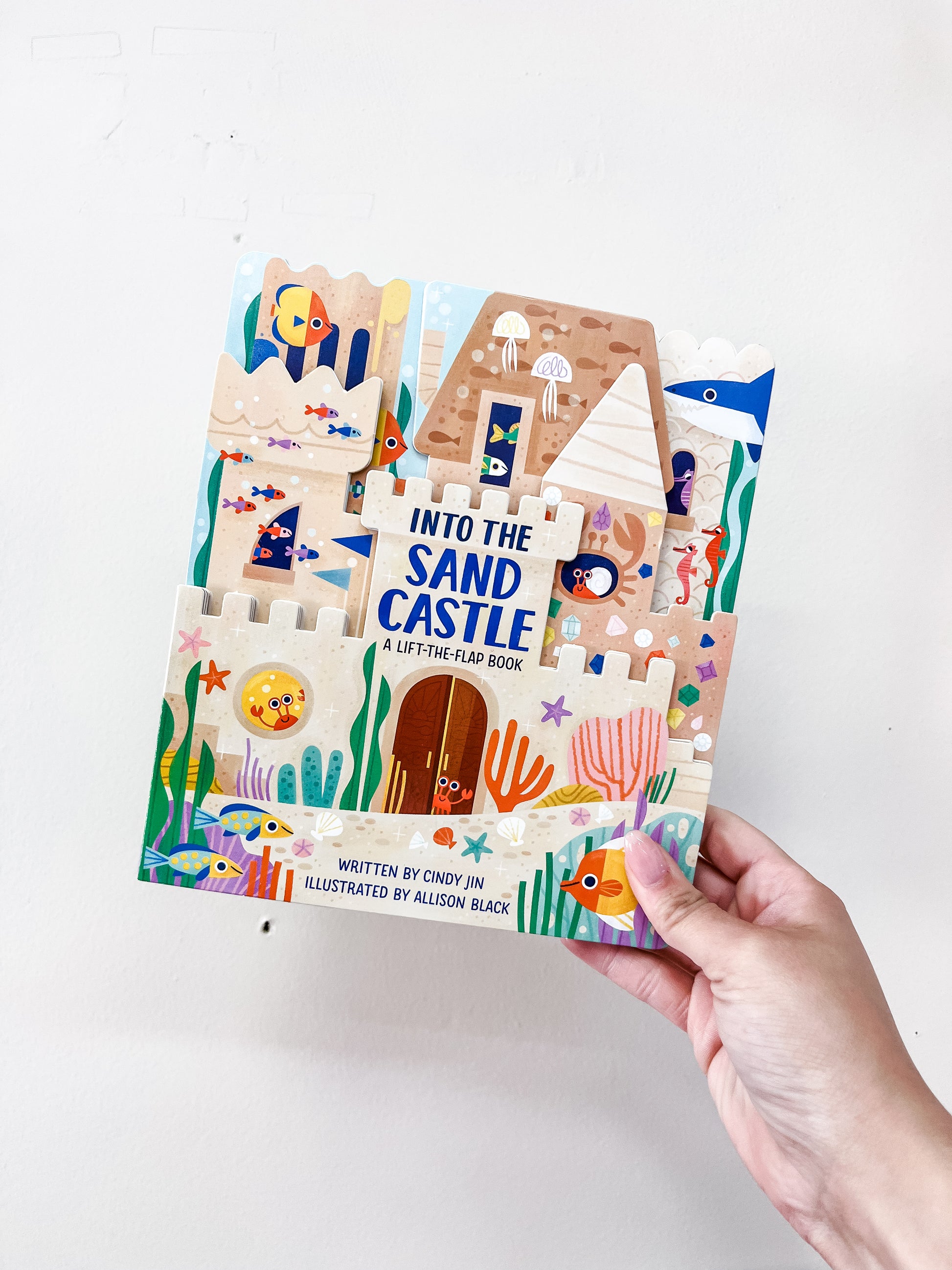 A hand holds the "Into the Sand Castle Book" by Simon & Schuster, featuring playful sea creatures, a sandcastle, coral, and a pyramid on the vibrant cover.