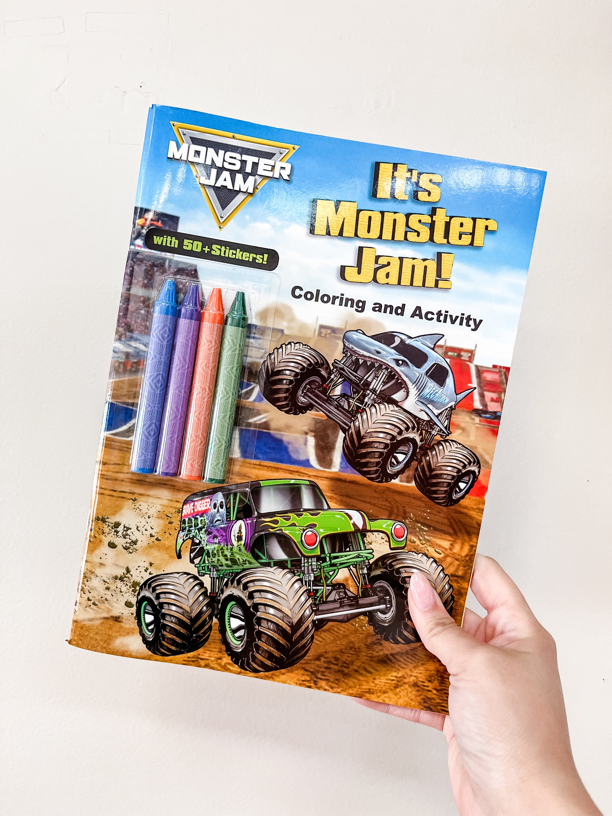 A hand holds the Simon & Schuster It's Monster Jam Book, which features two monster trucks, crayons, and "It's Monster Jam! 50+ stickers!" text on the colorful cover.