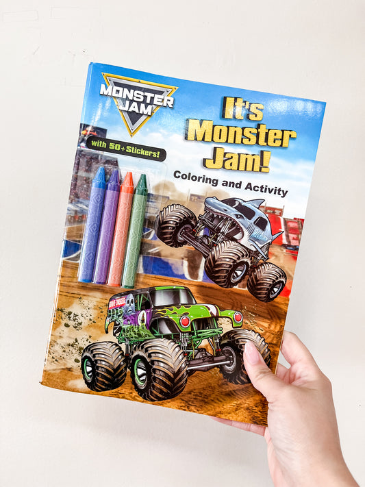 A hand holds the Simon & Schuster It's Monster Jam Book, which features two monster trucks, crayons, and "It's Monster Jam! 50+ stickers!" text on the colorful cover.