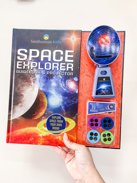A hand holds the Simon & Schuster Space Explorer Guide Book & Projector, featuring a galaxy-themed cover and projector discs for viewing 20 space-themed images.