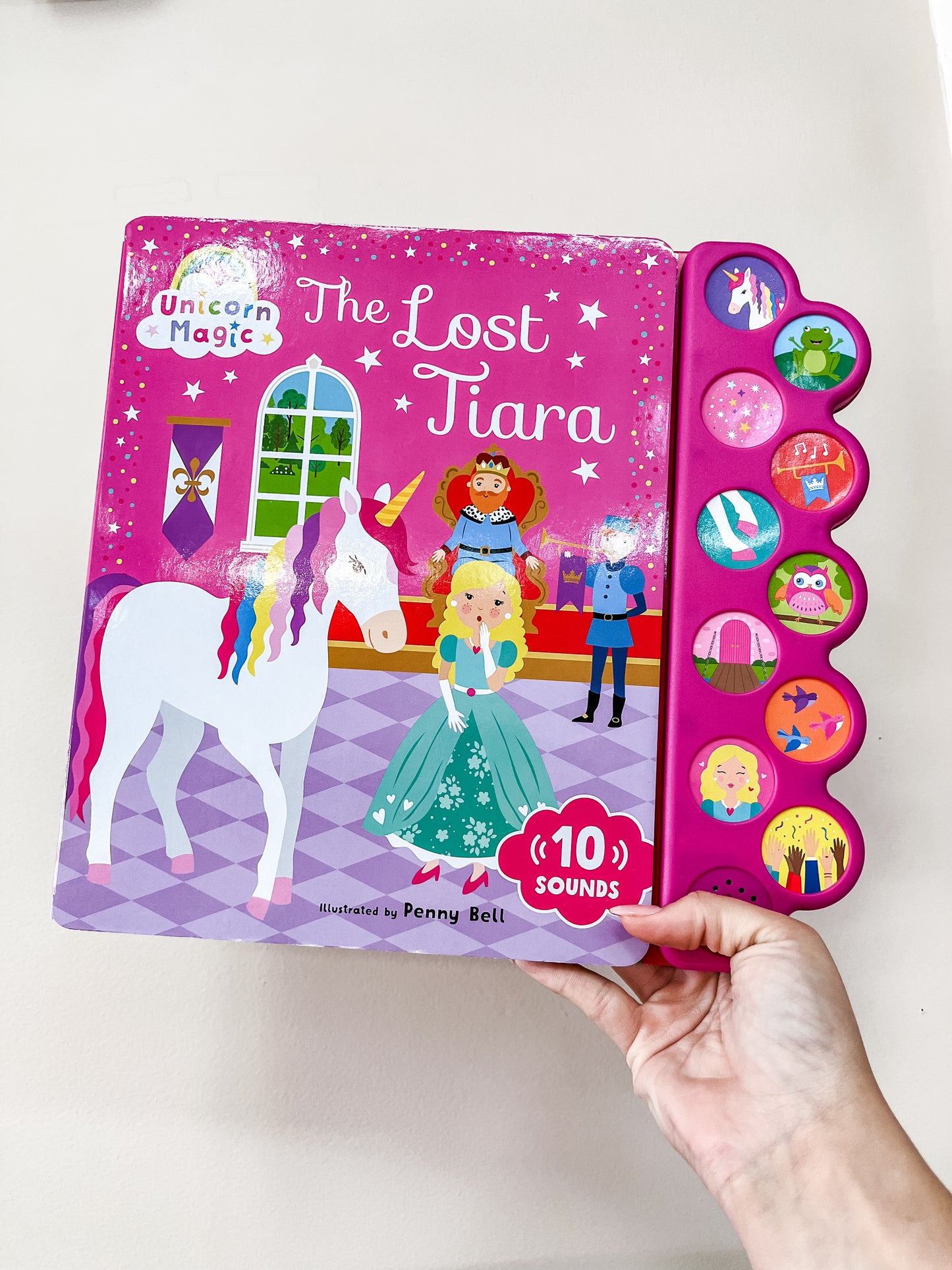A hand holds Unicorn Magic the Lost Tiara: Sound Book by Simon & Schuster. The cover shows a unicorn, princess, and prince, and the panel features sound buttons with colorful character illustrations.