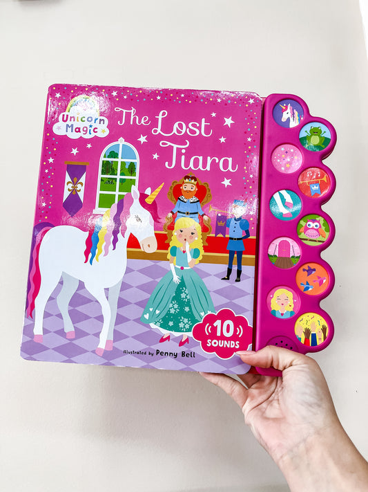 A hand holds Unicorn Magic the Lost Tiara: Sound Book by Simon & Schuster. The cover shows a unicorn, princess, and prince, and the panel features sound buttons with colorful character illustrations.
