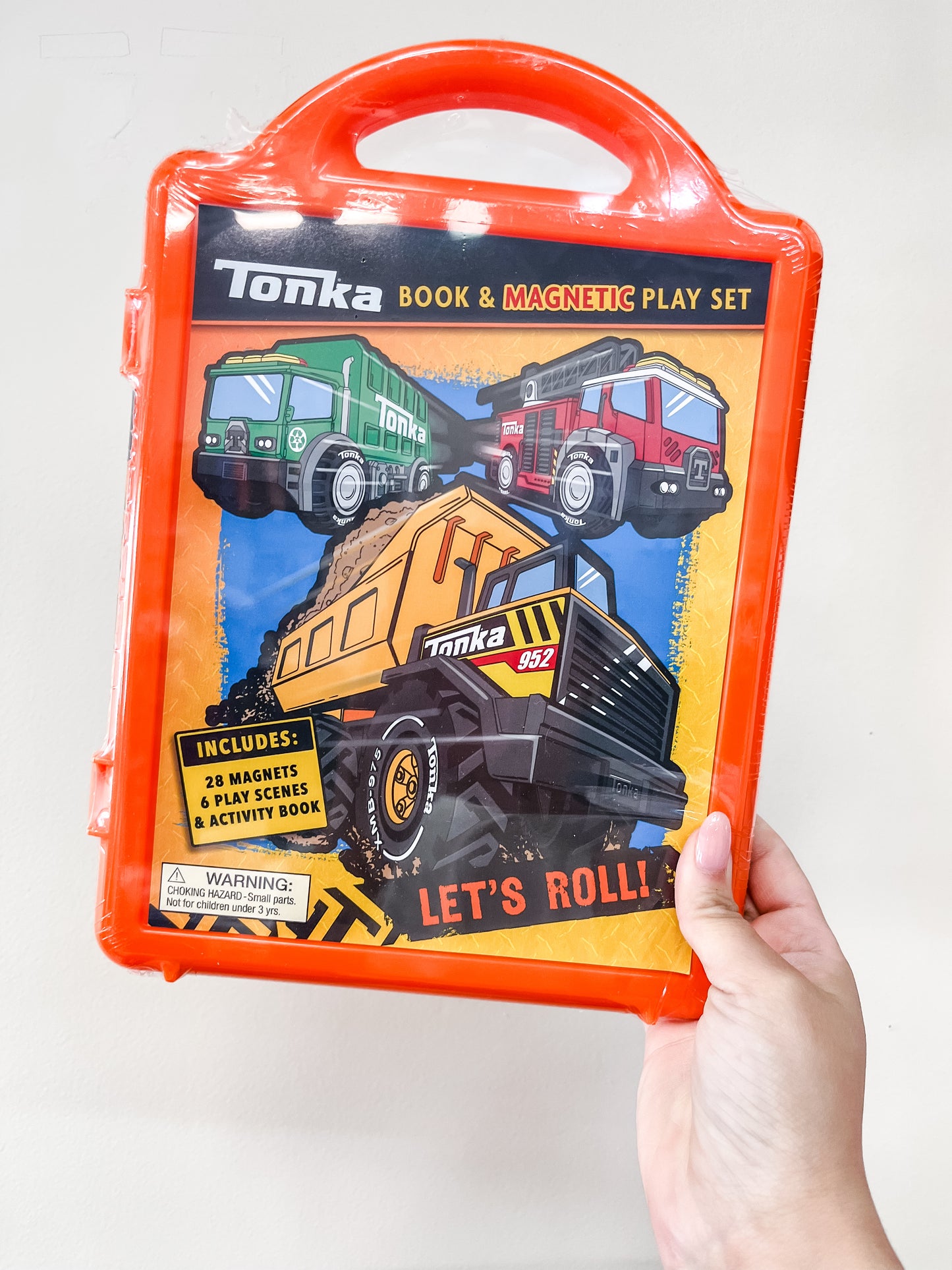 A hand holds the Simon & Schuster Tonka: Magnetic Play Set: Let's Roll Book in an orange case, featuring construction truck illustrations and the text "Let's Roll!" The set includes magnets, play scenes, and an activity book.