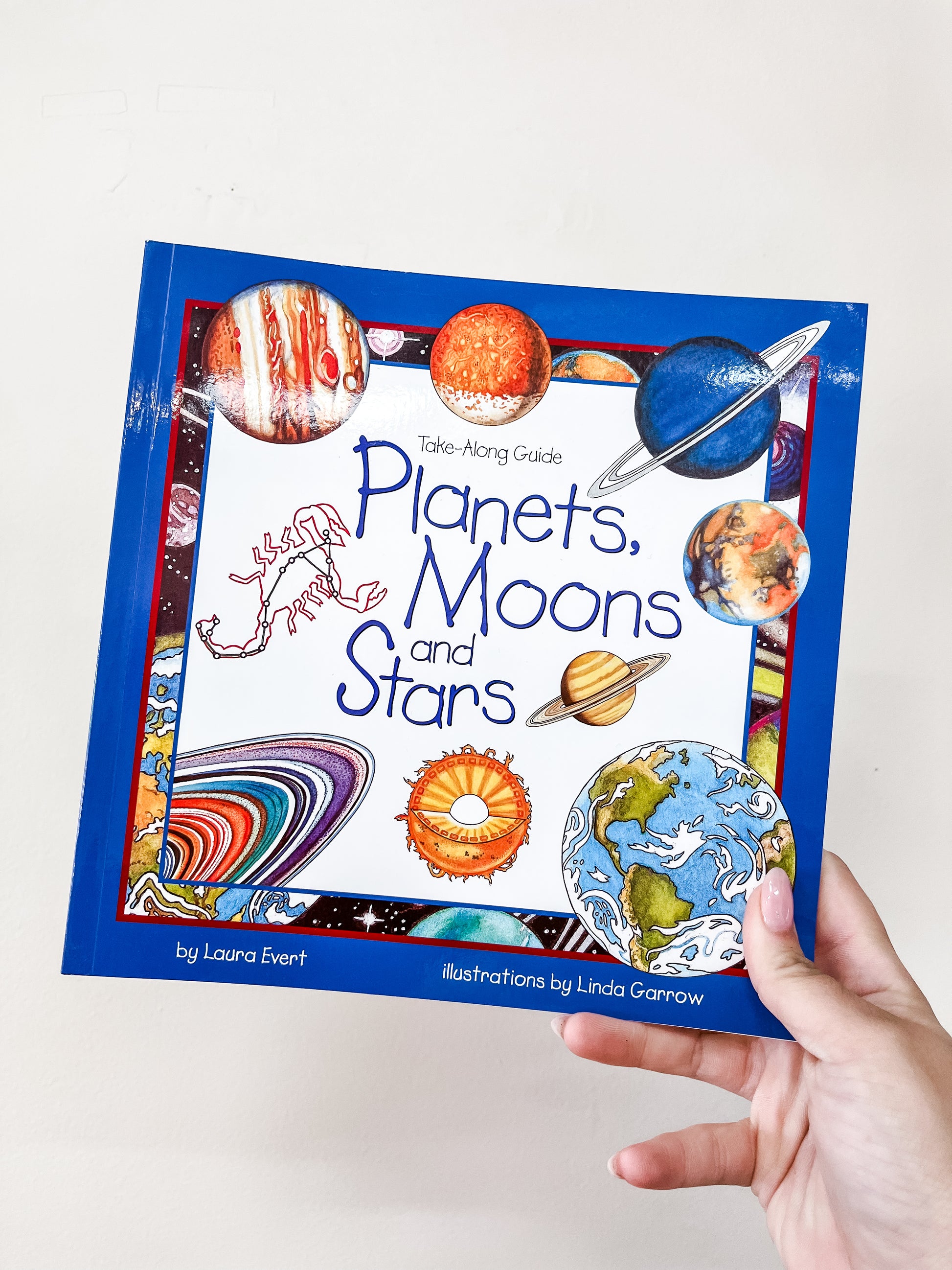 A hand holds up the Simon & Schuster Planets, Moons and Stars Book, which features colorful cover illustrations of planets, the Sun, Saturn, and constellations.