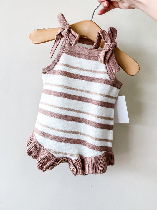 A hand holds a hanger displaying the Cocoa Cream Tie Bubble by Tiny Tot Bundles, a knitted baby romper with brown and white stripes, bow-tied shoulder straps, and ruffled leg openings.