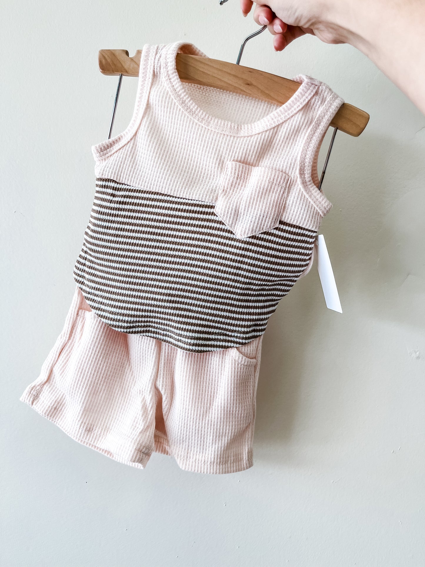 A hand holds Tiny Tot Bundles' Sandy Shores Two Piece—a sleeveless baby outfit with light pink ribbed fabric, striped top, chest pocket, and matching shorts—displayed against a plain light background.