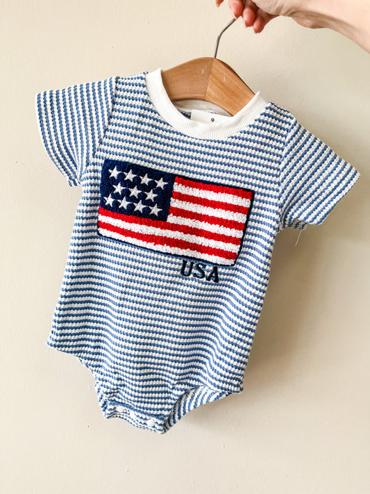 A hand holds the Tiny Tot Bundles Stars & Stripes Bubble, a blue and white striped baby romper on a wooden hanger, featuring a textured American flag and "USA" on the front.