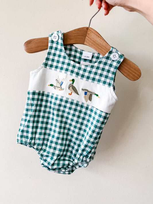 The Duck Pond Gingham Bubble by Tiny Tot Bundles features green and white checks, white shoulder straps, and embroidered flying ducks. Displayed on a wooden hanger against a plain wall.