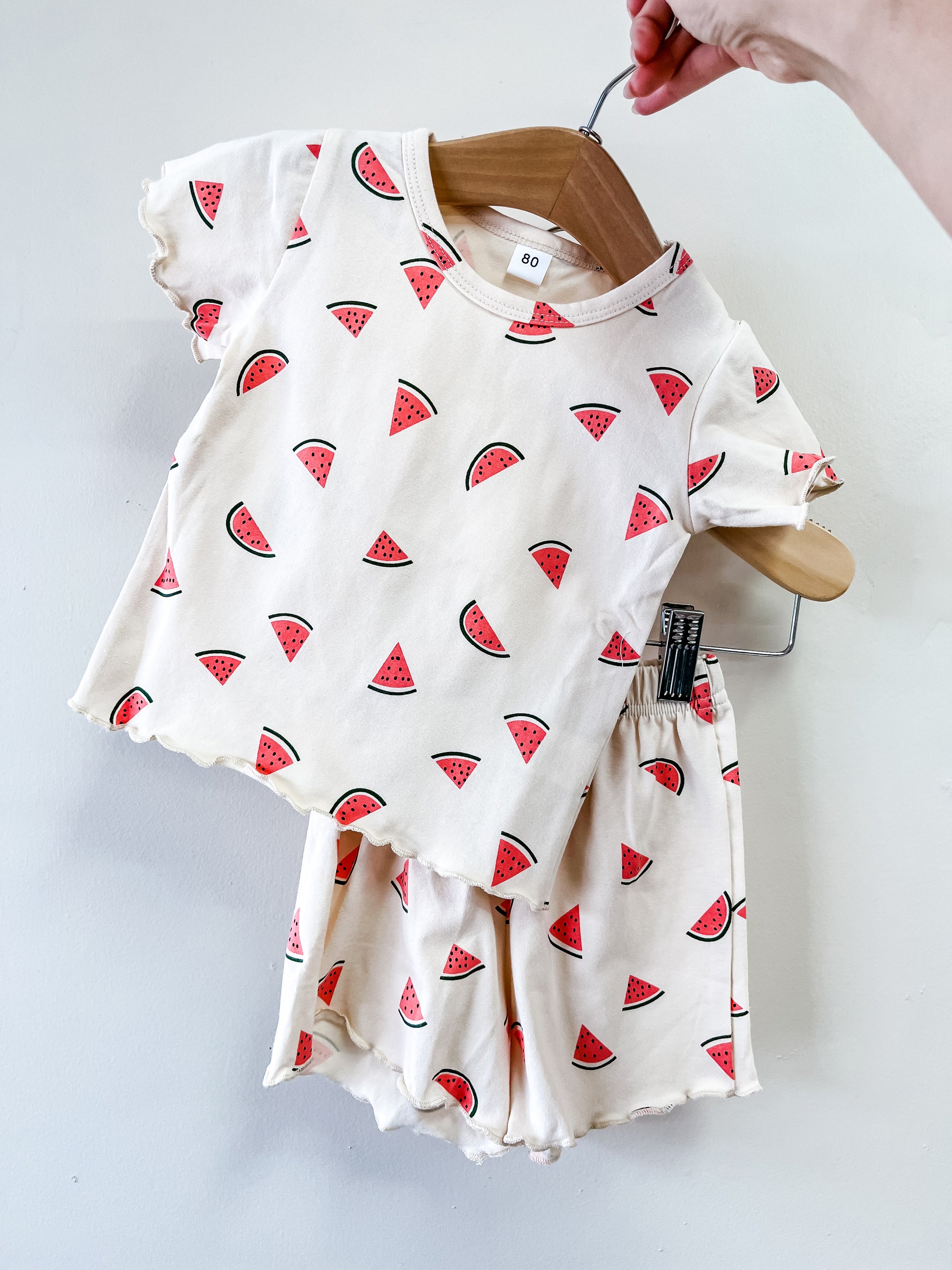 A hand holds Tiny Tot Bundles' Watermelon Dreams Set: a matching toddler outfit with a cream-colored short-sleeve shirt and shorts, both adorned with a playful watermelon slice print.