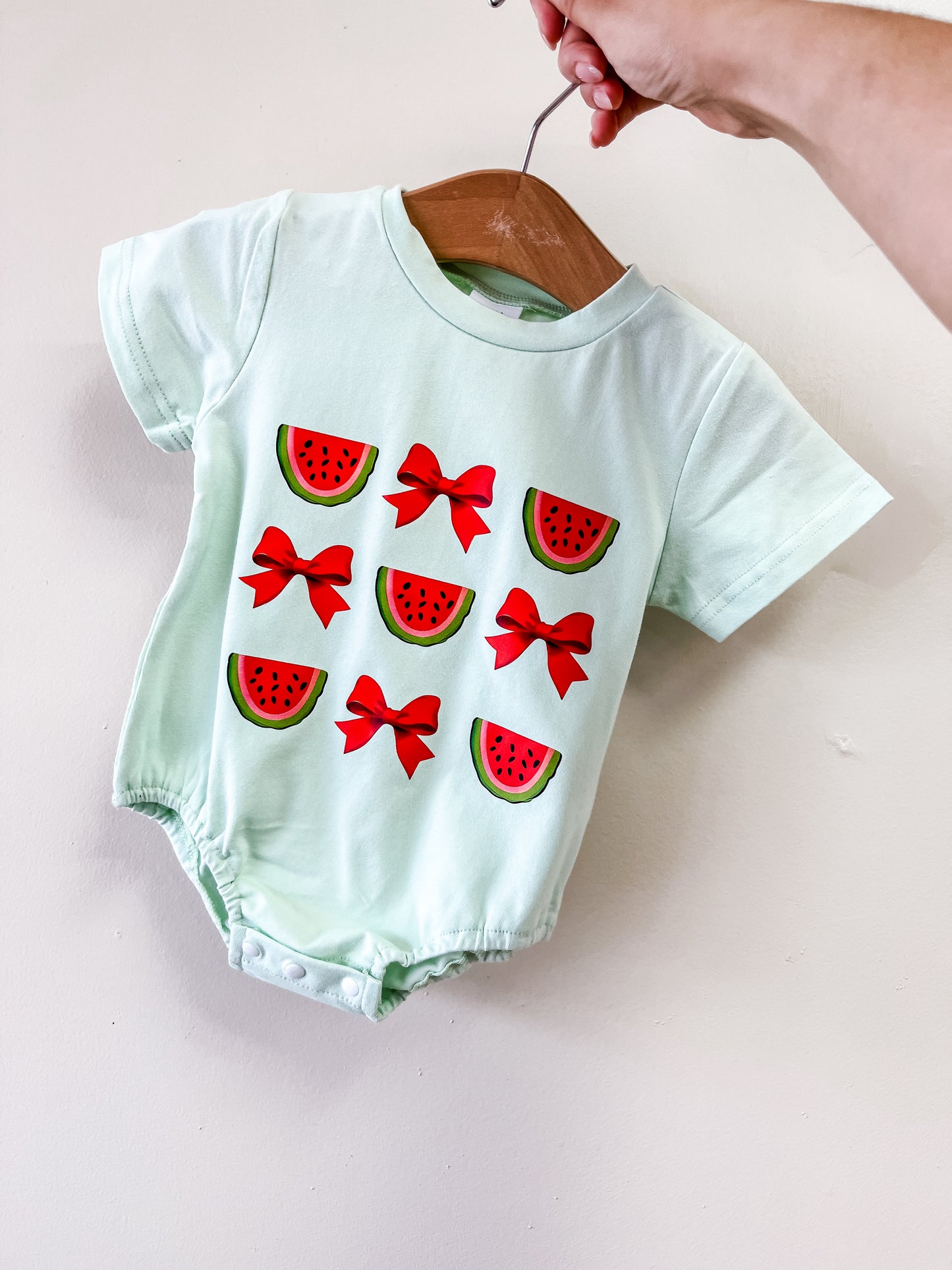 A hand holds Tiny Tot Bundles' Watermelon Bow Bubble on a wooden hanger. The light green onesie is decorated with six watermelon slices and three red bows on the front.