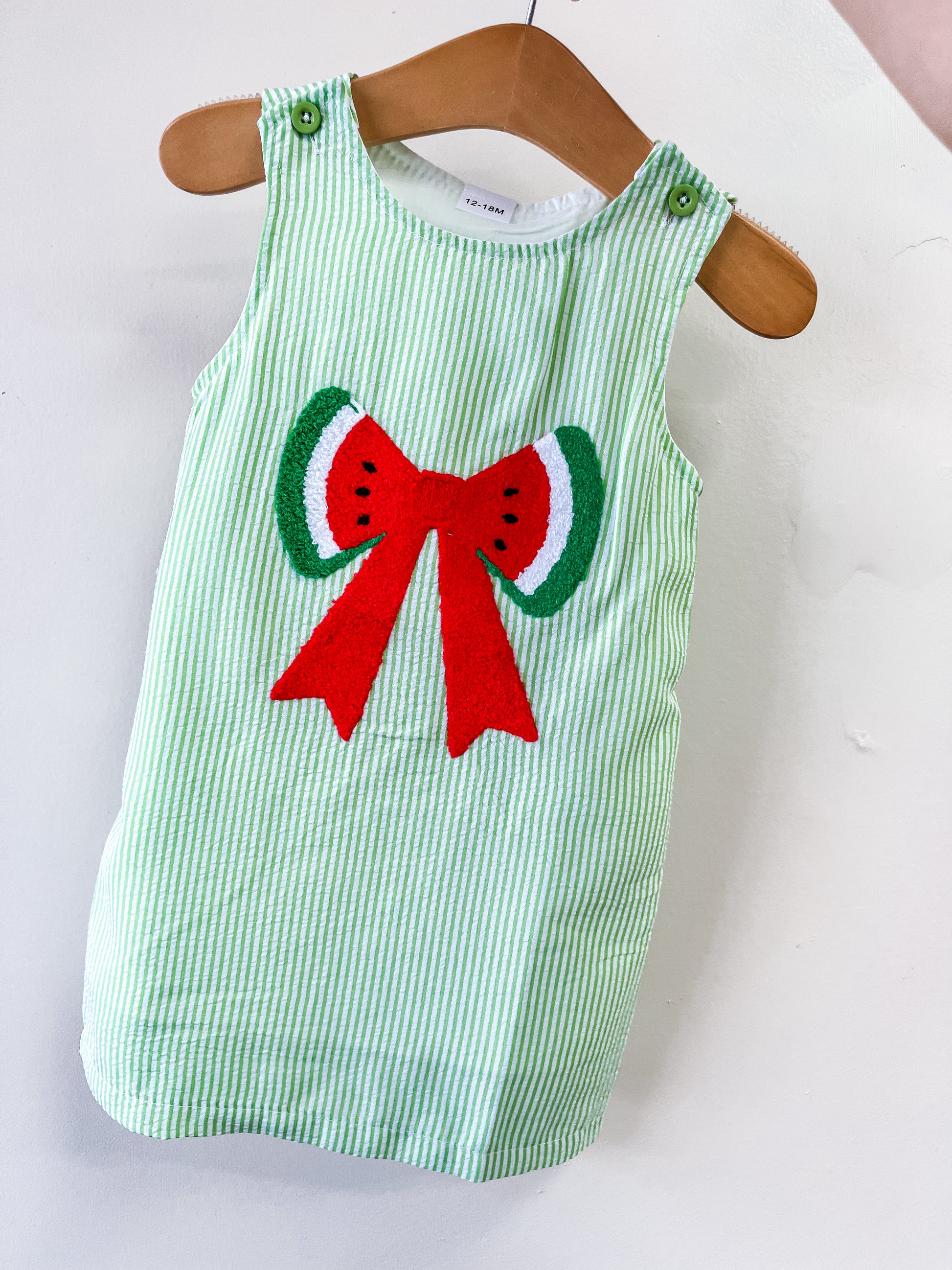 The Tiny Tot Bundles Watermelon Wishes Dress is a green and white striped sleeveless toddler dress featuring an embroidered red bow with watermelon slice designs on the front, displayed on a wooden hanger.