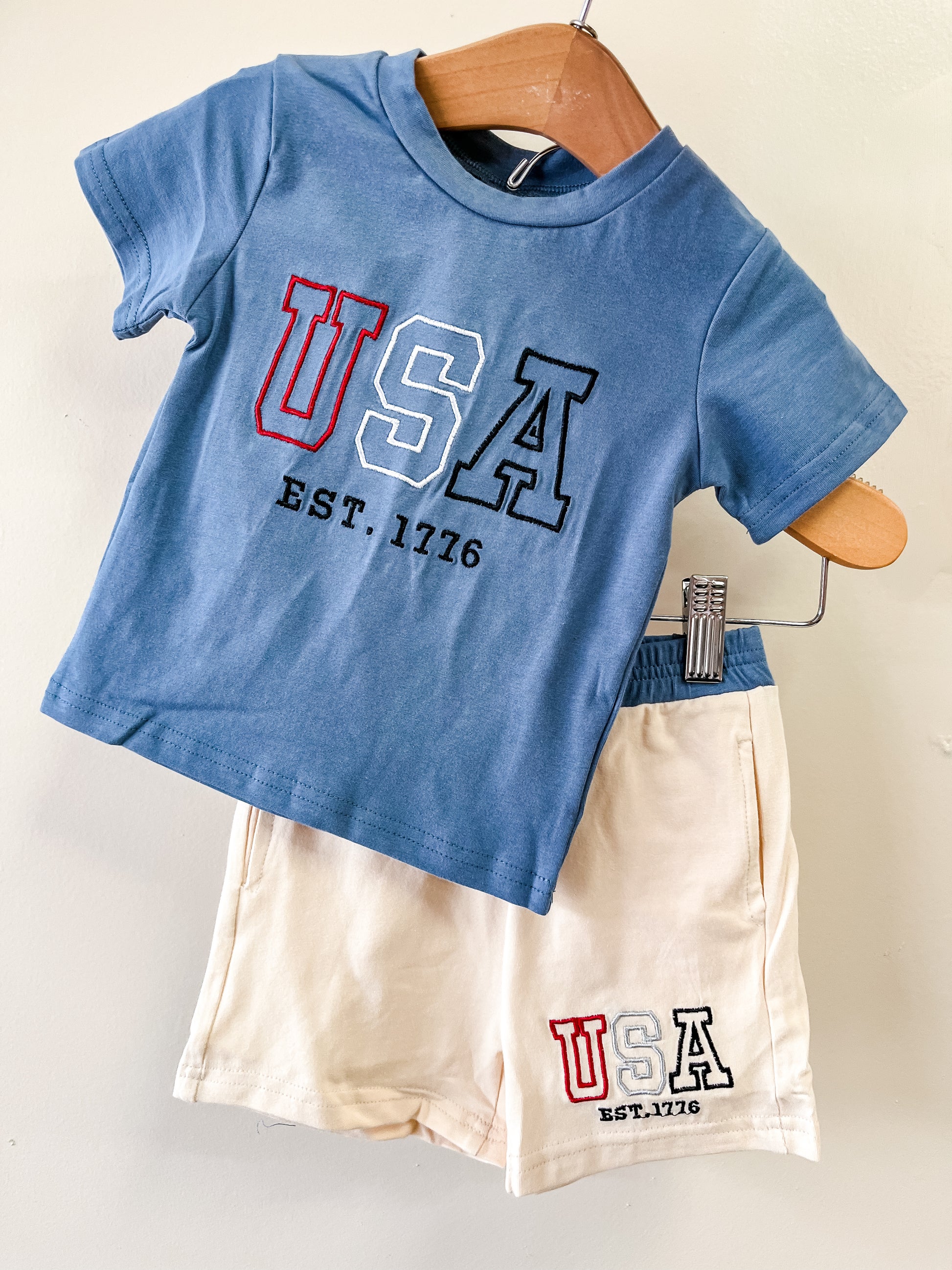 The Little Patriot Set by Tiny Tot Bundles features a blue tee and cream shorts, both with USA EST. 1776 designs, displayed together on a wooden hanger against a light background.