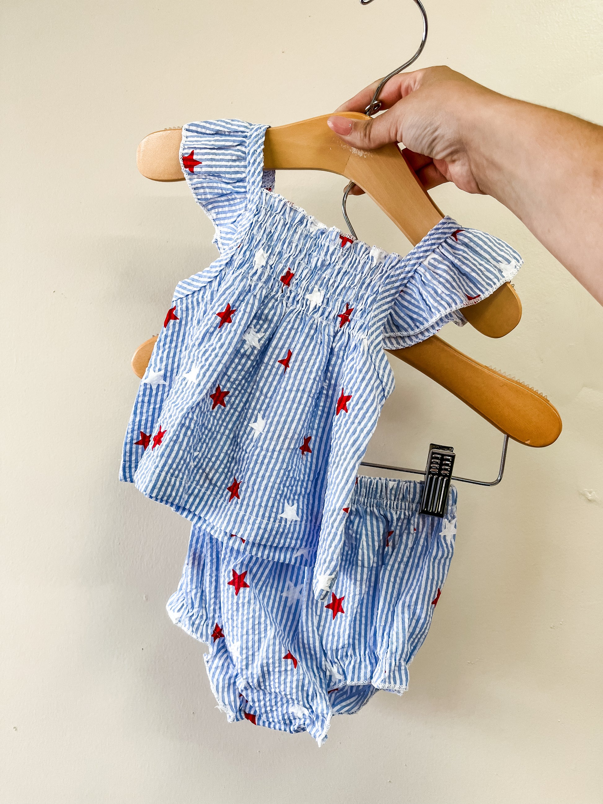 A hand displays the Tiny Tot Bundles Americana Babe Set—a blue and white striped toddler outfit with red stars, featuring a ruffled sleeveless top and matching bloomers—against a plain light wall.