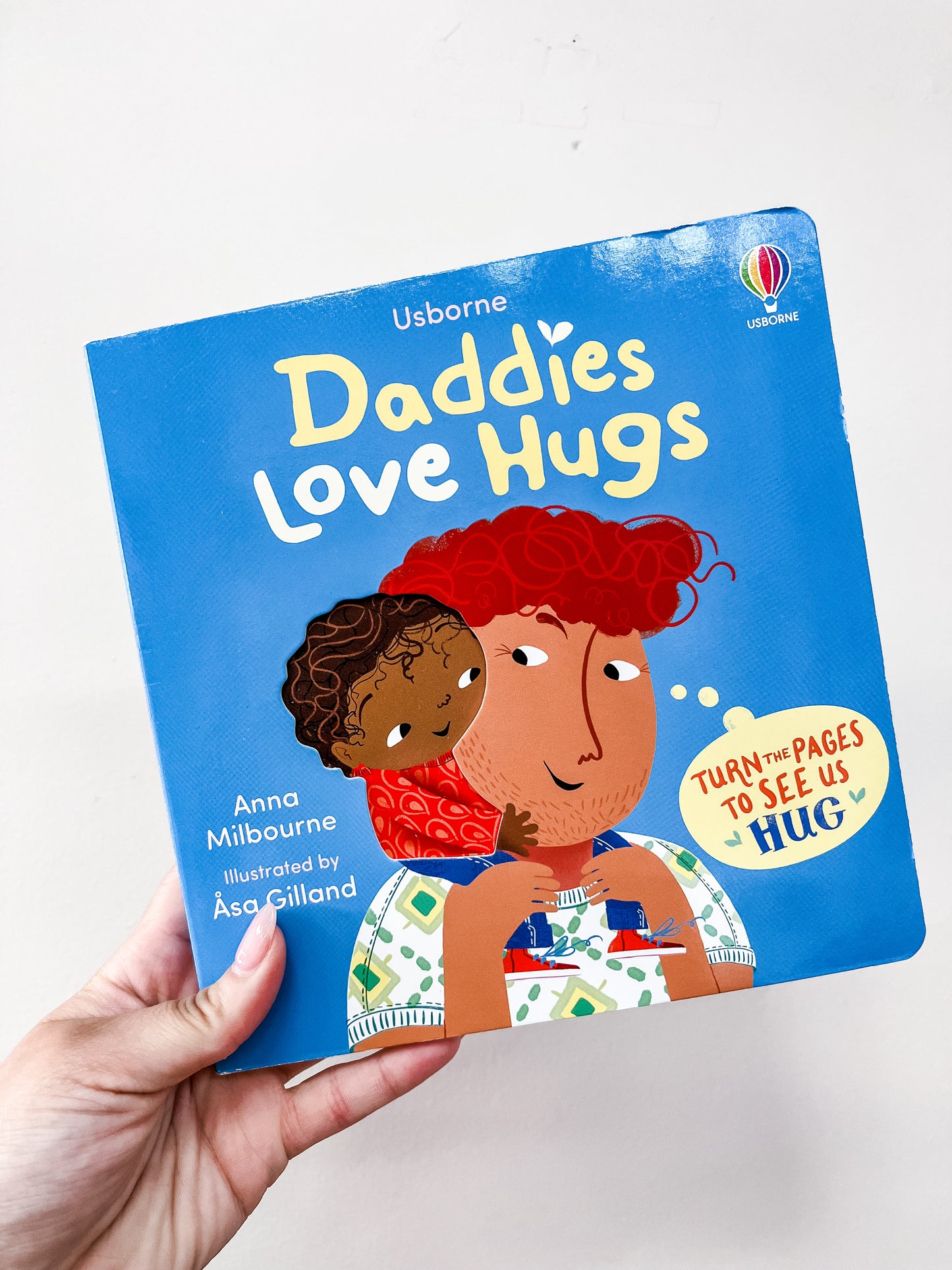 A hand holds the Daddies Love Hugs Board Book from Tiny Tot Bundles. The cover features a daddy hugging a child against a blue background.