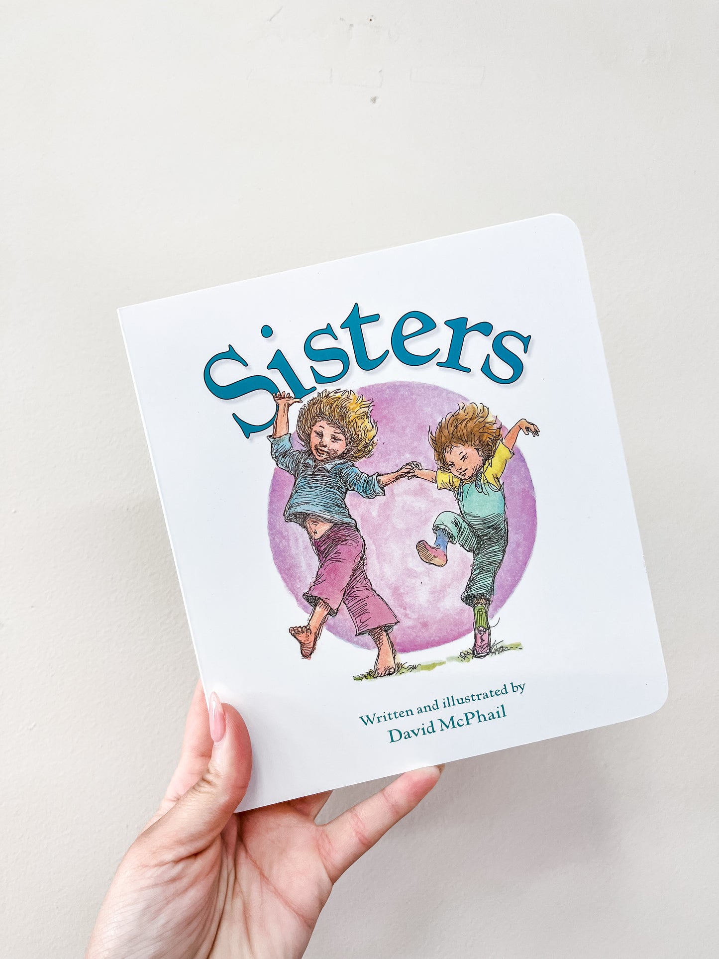 A hand holds the Sisters Book from Harper Collins, with a cover showing two smiling girls dancing in front of a purple circle.