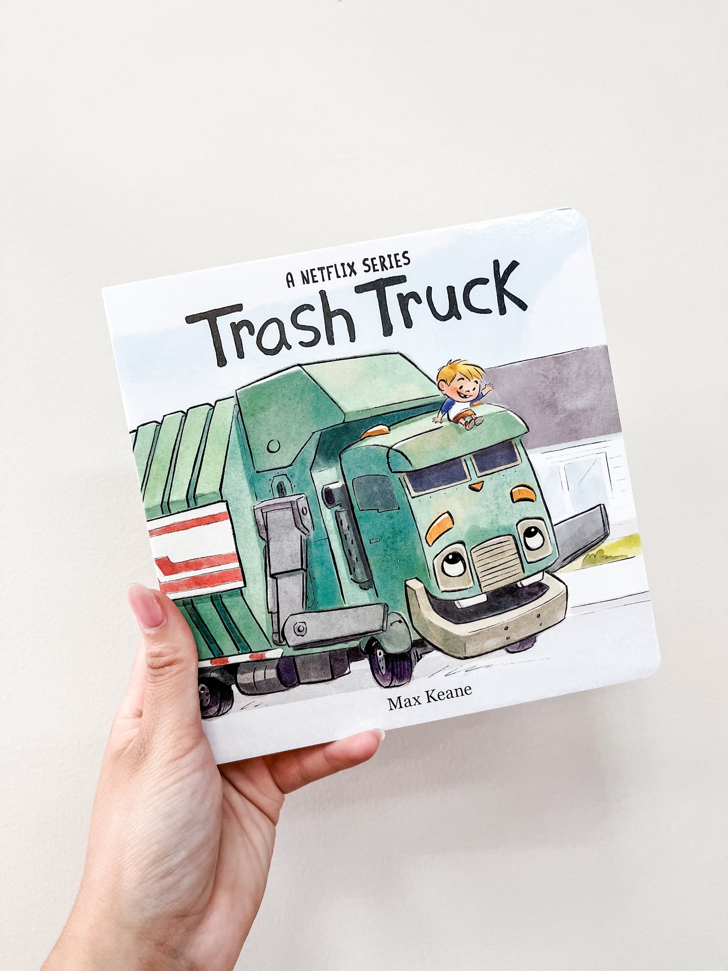 A hand holds the "Trash Truck Board Book" by Harper Collins, showing a smiling boy riding atop a friendly green trash truck. The white cover features simple, colorful artwork perfect for children.