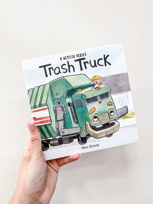 A hand holds the "Trash Truck Board Book" by Harper Collins, showing a smiling boy riding atop a friendly green trash truck. The white cover features simple, colorful artwork perfect for children.