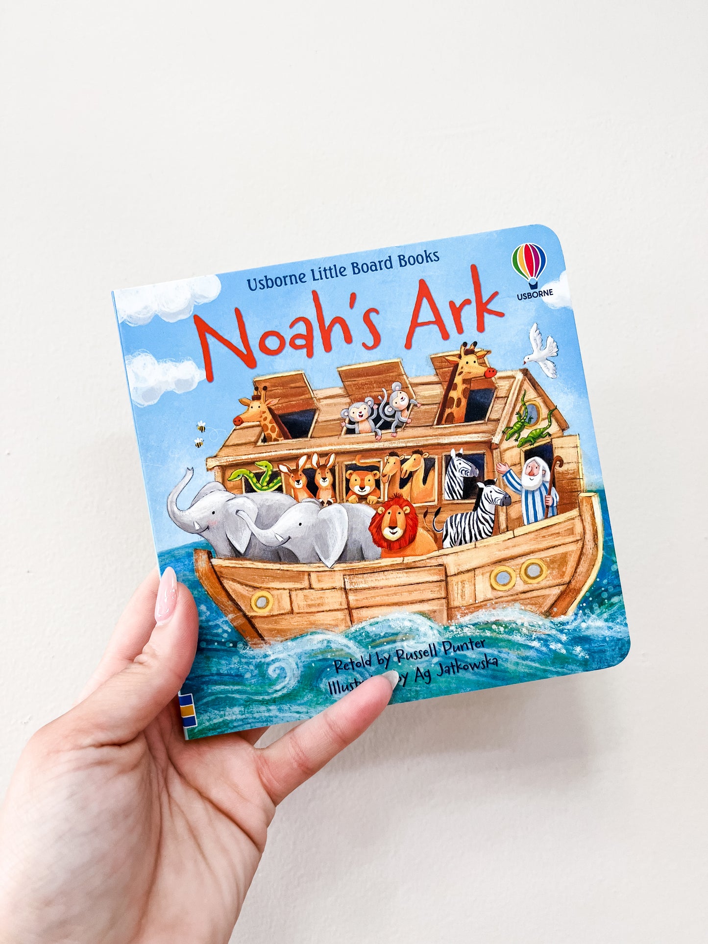 A hand holds the Noah's Ark Book by harper Collins, a colorful children's board book with illustrated animals—including elephants, lions, and zebras—aboard a wooden ark under a blue sky.