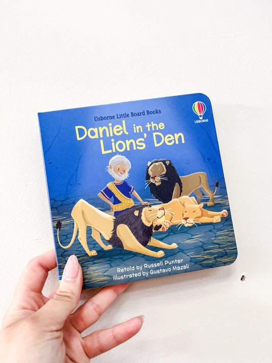 A hand holds the "Daniel in the Lions' Den Board Book" by Harper Collins, featuring an illustration of Daniel with three lions on the cover.