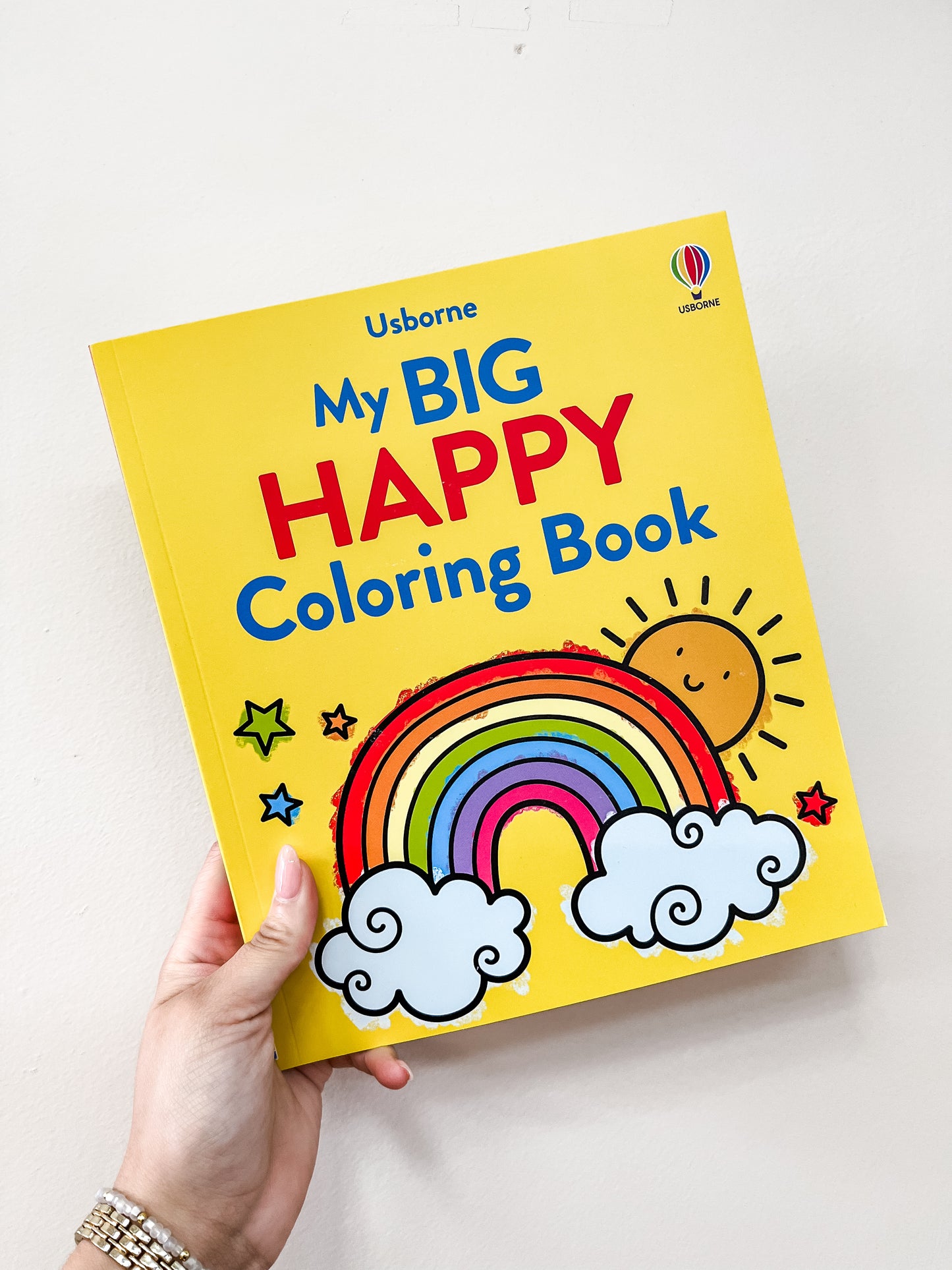 A hand holds the harper Collins My Big Happy Coloring Book, featuring a yellow cover with a rainbow, sun, stars, and clouds.
