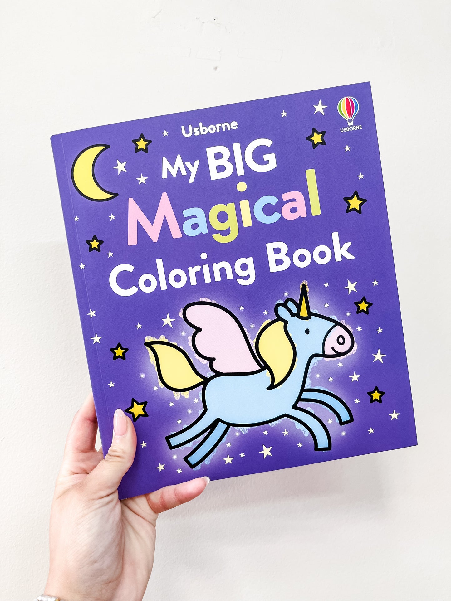 A hand holds a purple book titled My Big Magical Coloring Book by harper Collins, featuring a winged unicorn, stars, and a crescent moon on the cover.