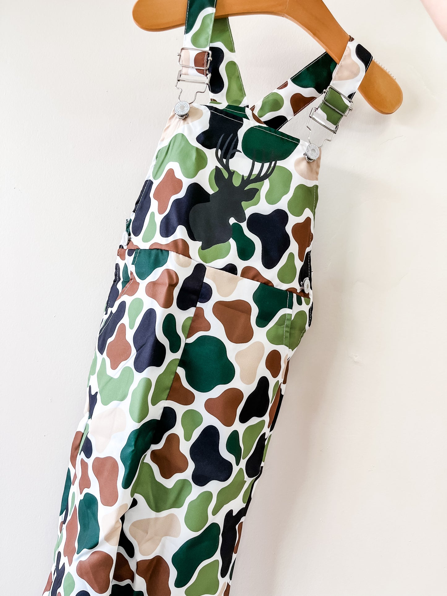Tiny Tot Bundles Camo Overalls feature green, brown, tan, and black camouflage with a black deer head silhouette on the chest, displayed on a wooden hanger against a white wall.