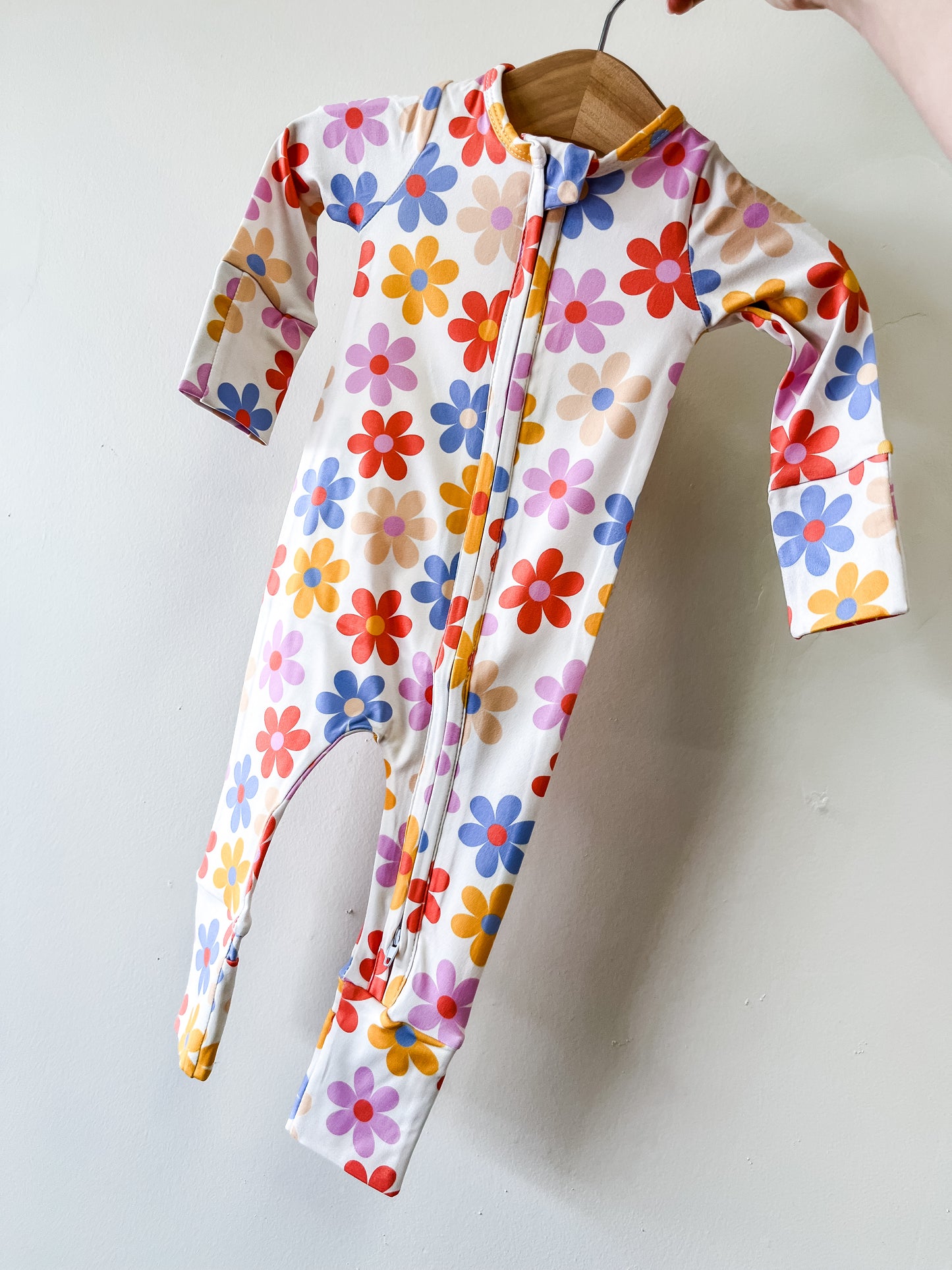 The Tiny Tot Bundles Spring Floral Zippy, a long-sleeved baby romper with a colorful red, yellow, blue, and purple floral pattern, is shown on a small wooden hanger against a plain white background.