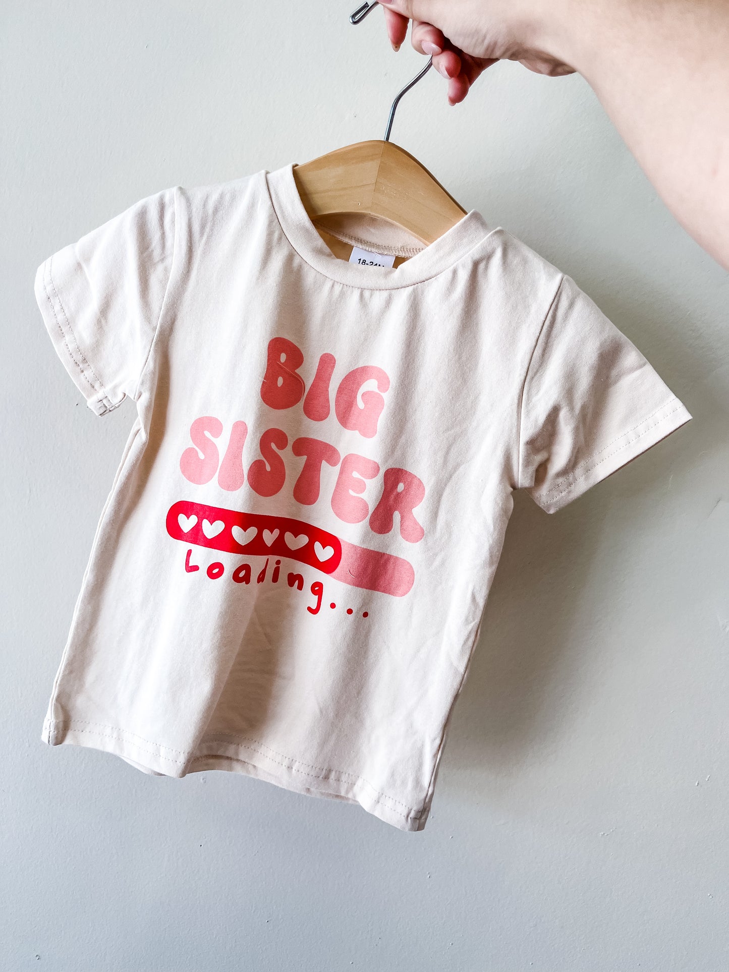 A hand holds the Tiny Tot Bundles Big Sister Loading Tee, a small white T-shirt on a hanger with pink "BIG SISTER" text above “Loading…” and a heart-filled progress bar.