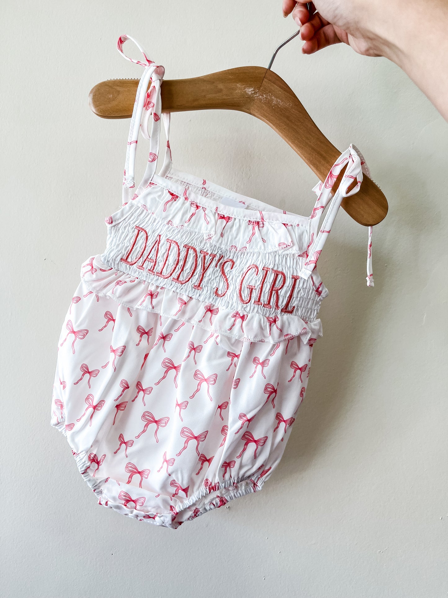 A hand holds a wooden hanger with the Tiny Tot Bundles Daddy's Girl Bow Romper, featuring pink bows, "DADDYS GIRL" embroidered in pink on the chest, tie straps, and a gathered waist and legs.