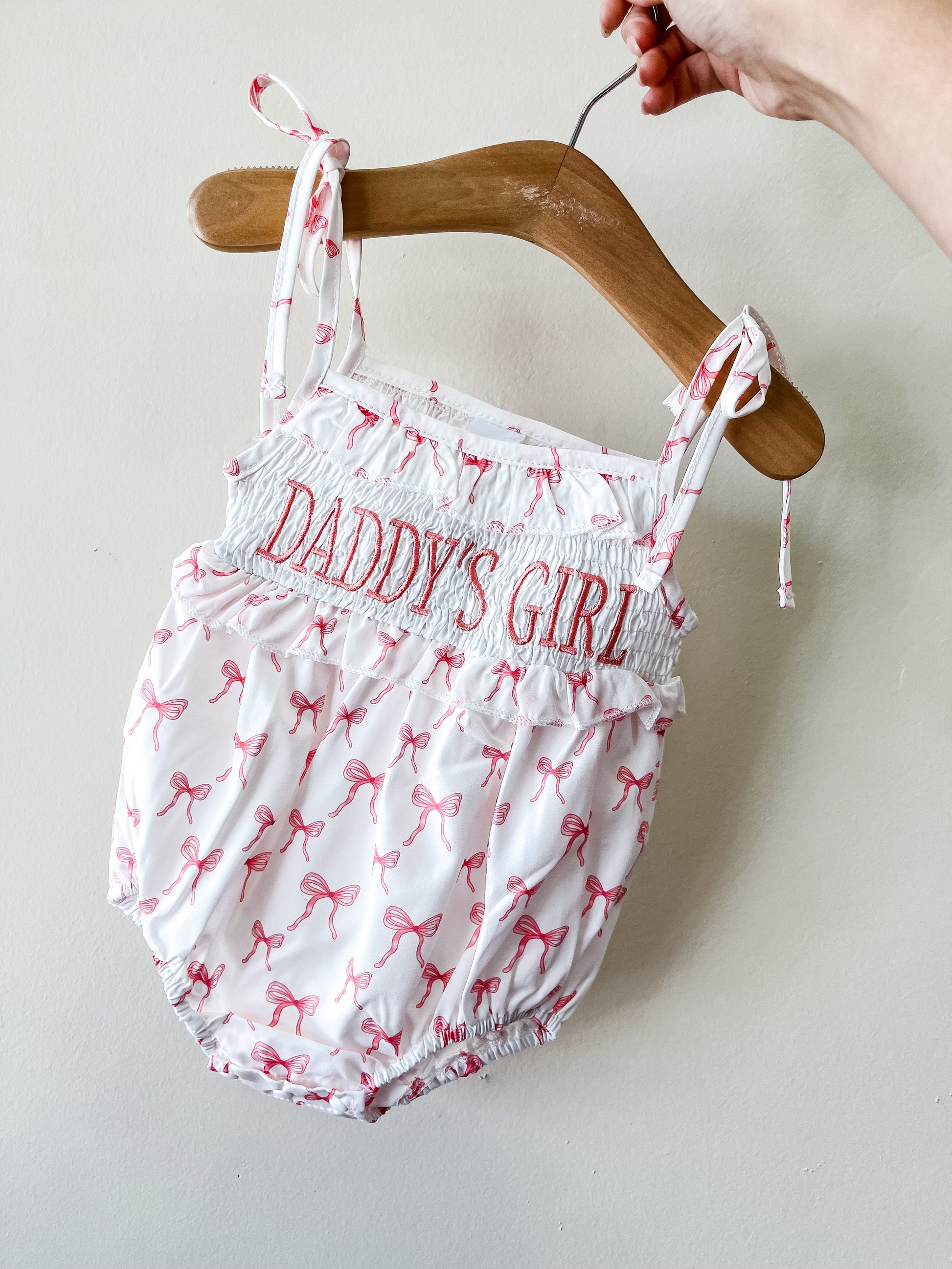 A hand holds a wooden hanger with the Tiny Tot Bundles Daddy's Girl Bow Romper, featuring pink bows, "DADDYS GIRL" embroidered in pink on the chest, tie straps, and a gathered waist and legs.