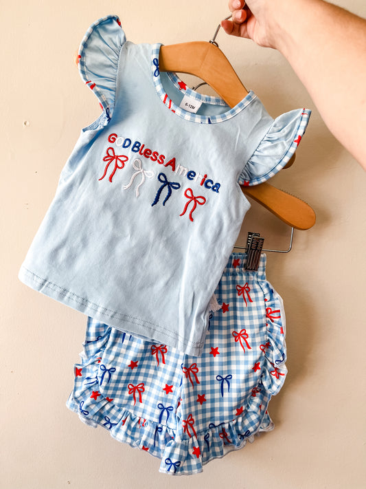 The Little Miss America Set by Tiny Tot Bundles features a light blue ruffled sleeve top with "God Bless America" and matching ruffled shorts, both adorned with red, white, and blue bows and stars. Displayed on wooden hangers against a beige wall.